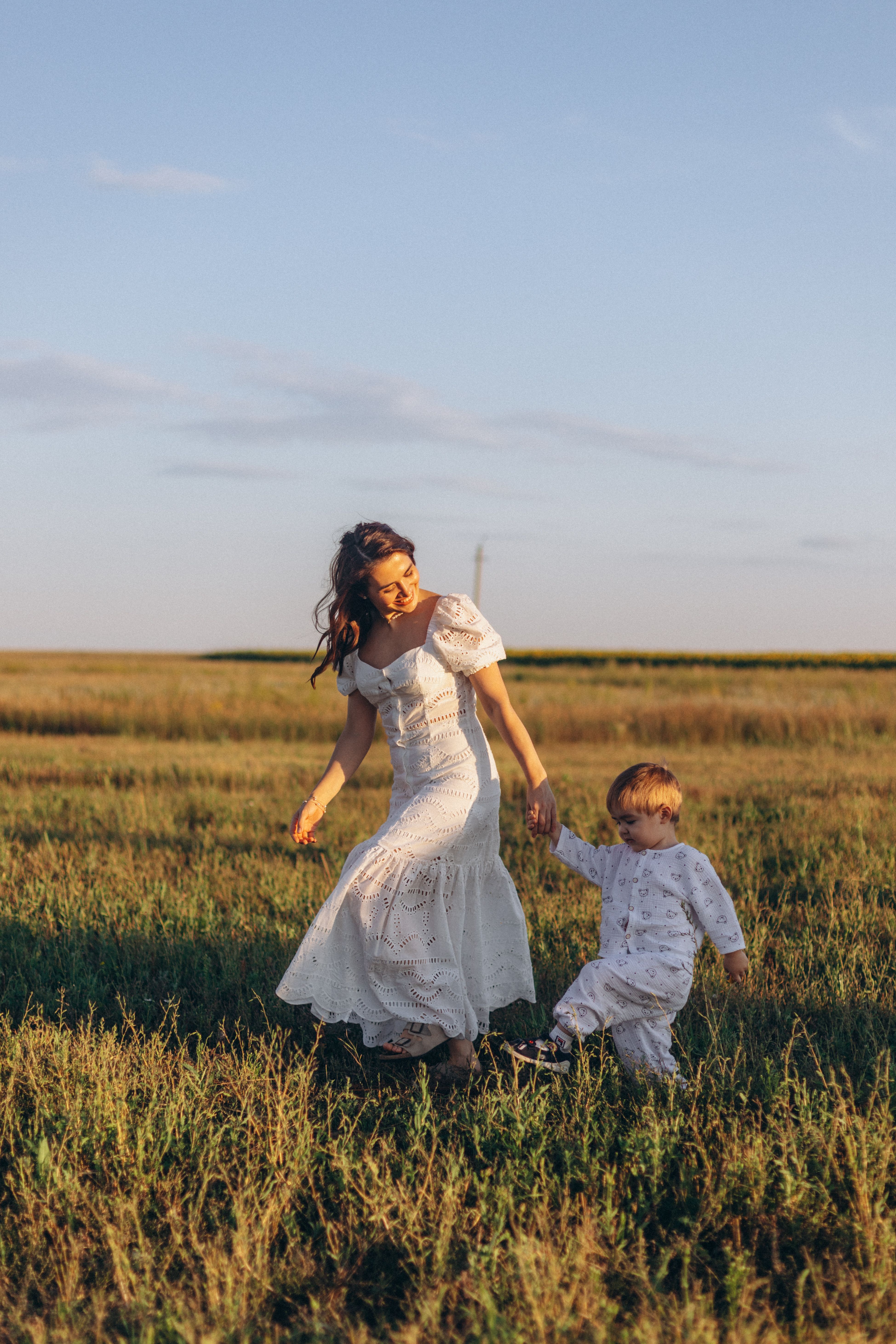 A mother and her young child walk hand in hand through a sunny field, capturing a warm and joyful moment.