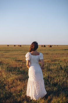 A woman in a white dress stands in an open field during sunset, capturing a serene rural scene.