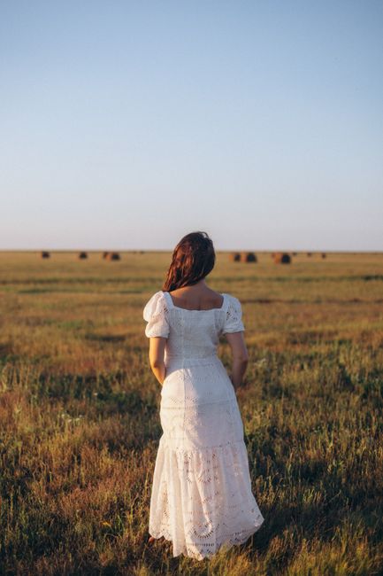 A woman in a white dress stands in an open field during sunset, capturing a serene rural scene.