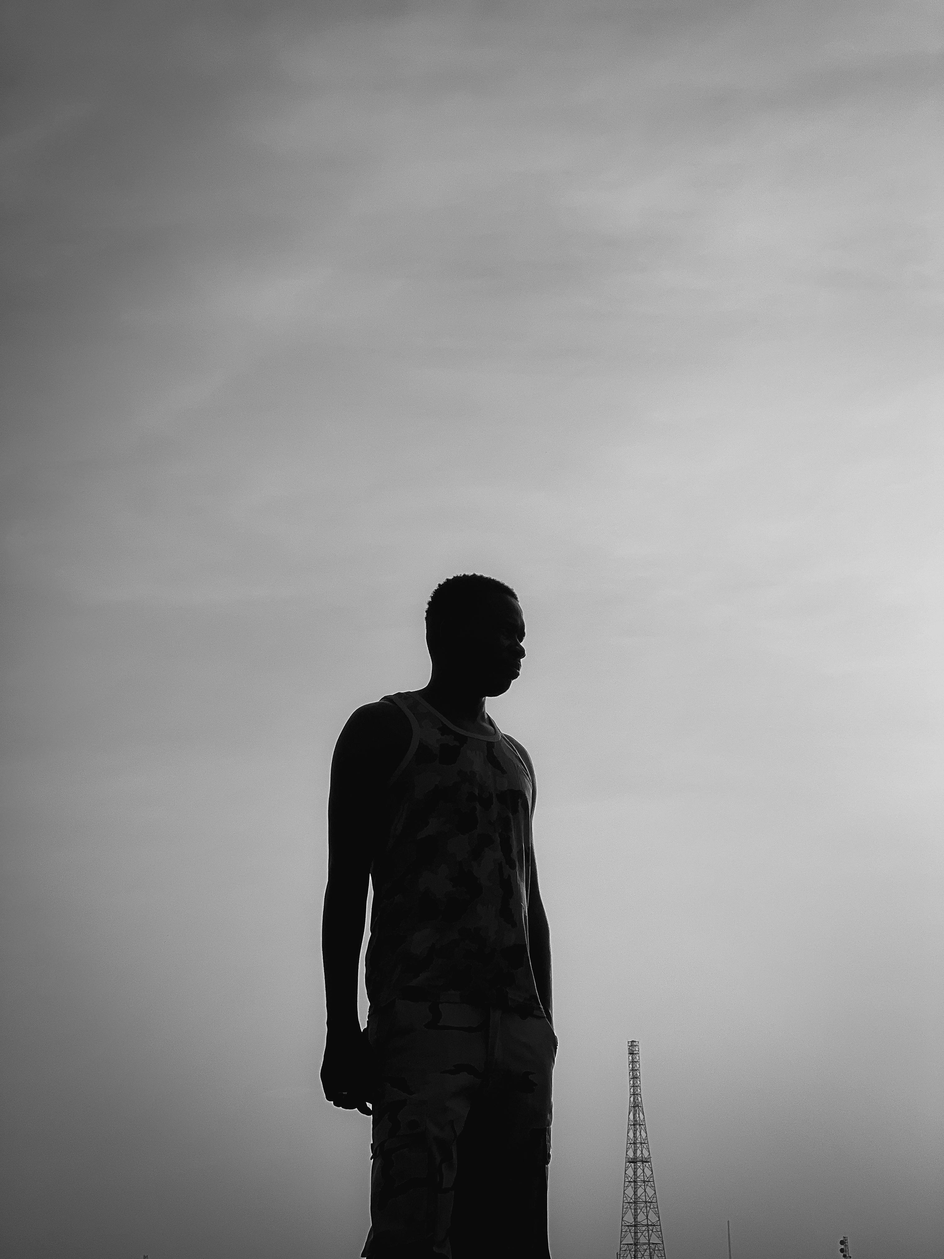 Free Black and white silhouette of a man standing outdoors near a tower during sunset. Stock Photo