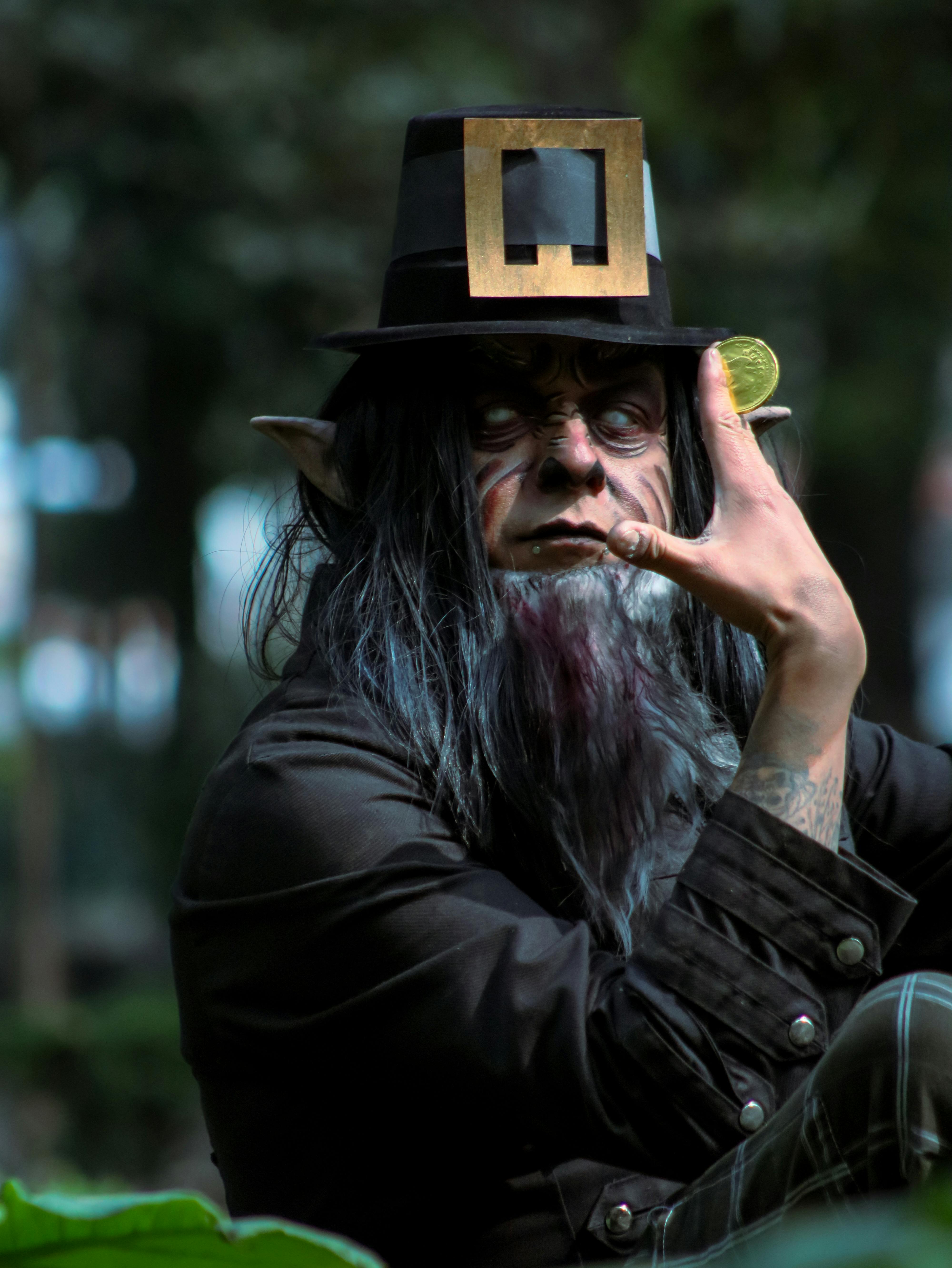 A man dressed as an elf holding a coin in a forested area, evoking a mystical atmosphere.