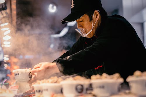 Man in Black Jacket and Black Hat Holding White Plastic Cups With Food