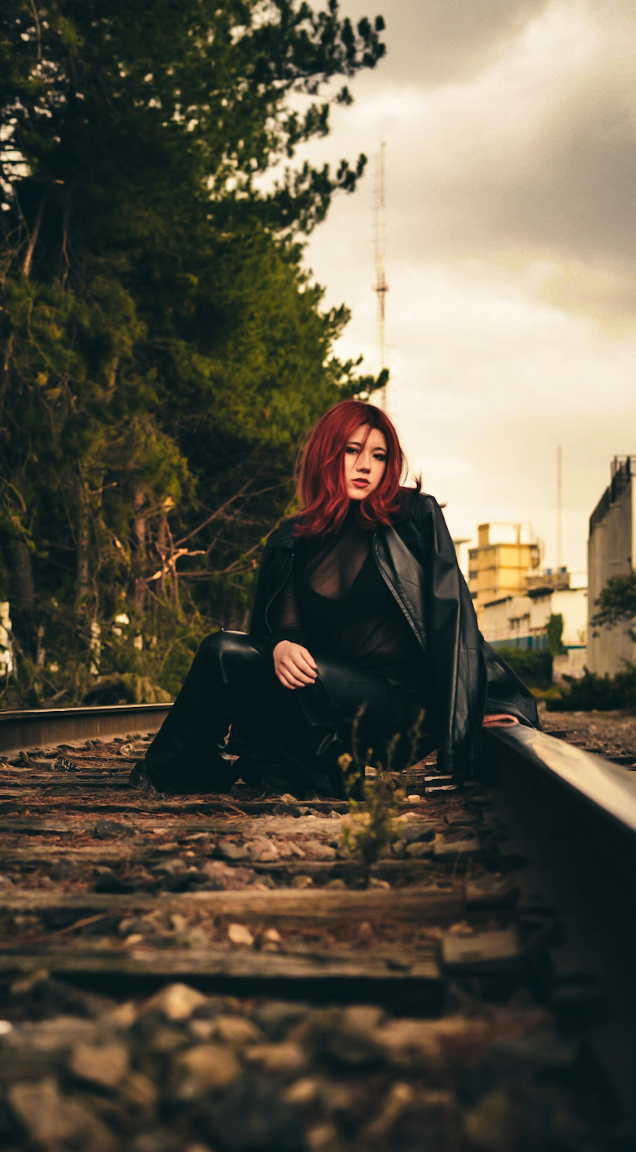 Gratis Una donna alla moda con i capelli rossi seduta sui binari del treno in un contesto urbano durante il giorno. Foto a disposizione