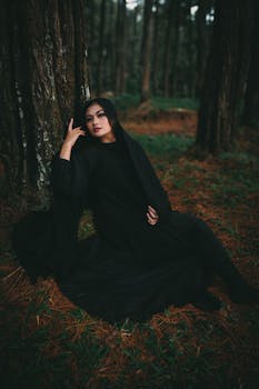 Woman in a dark cloak seated by a tree in a moody forest setting.