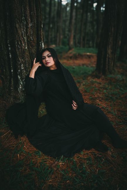 Woman in a dark cloak seated by a tree in a moody forest setting.