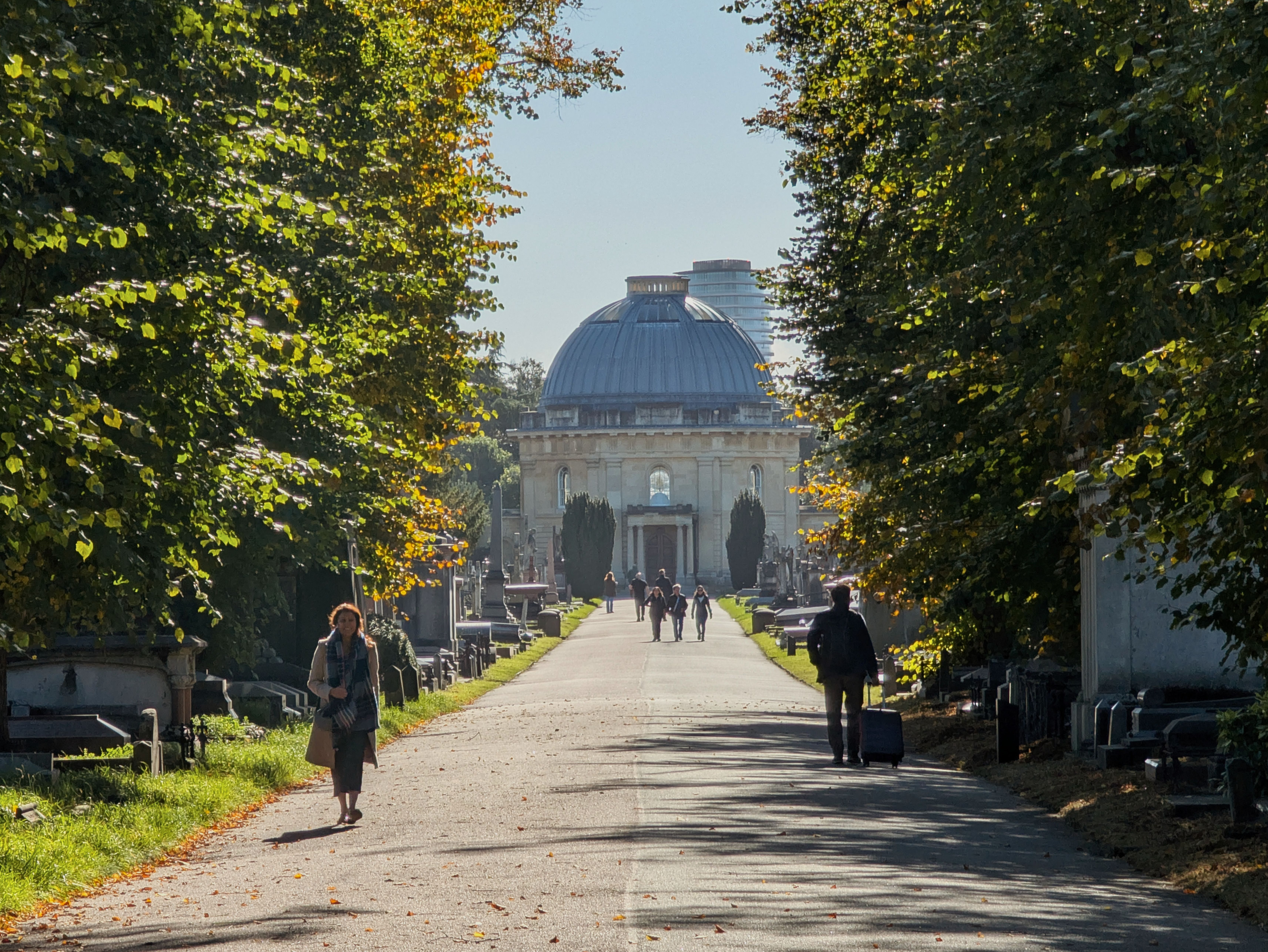 Victorian cemetery in London with visitors strolling along tree-lined path towards historical mausoleum.