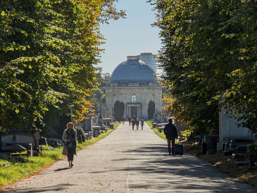 Victorian cemetery in London with visitors strolling along tree-lined path towards historical mausoleum.