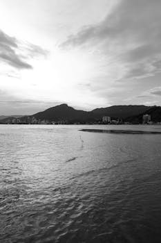 Black and white photo of a serene beach with distant mountains under a dramatic sky.