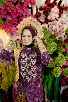 Smiling Indonesian bride in traditional dress with vibrant floral background. - Fotografer Sidoarjo