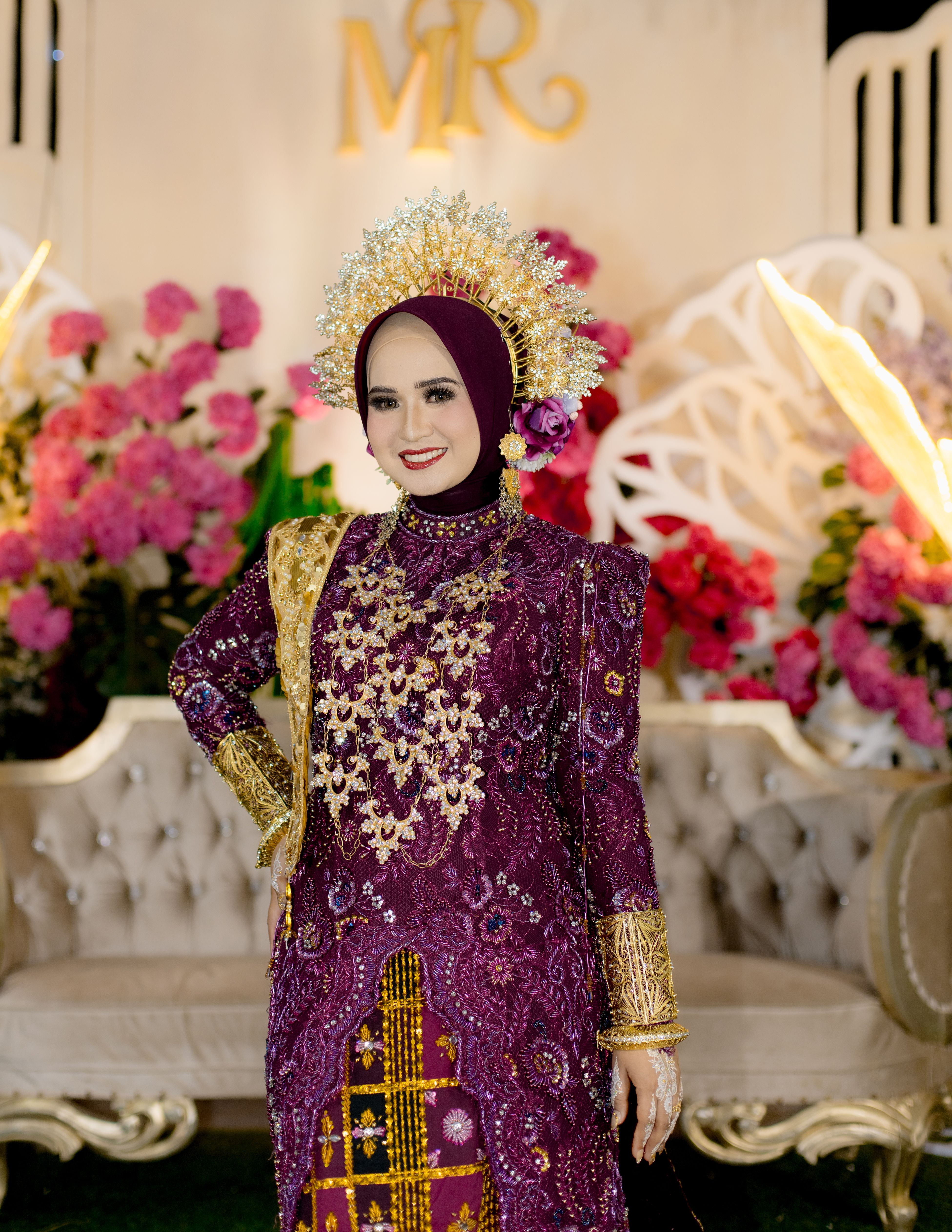 Elegant Indonesian bride in ornate purple bridal attire with floral backdrop.