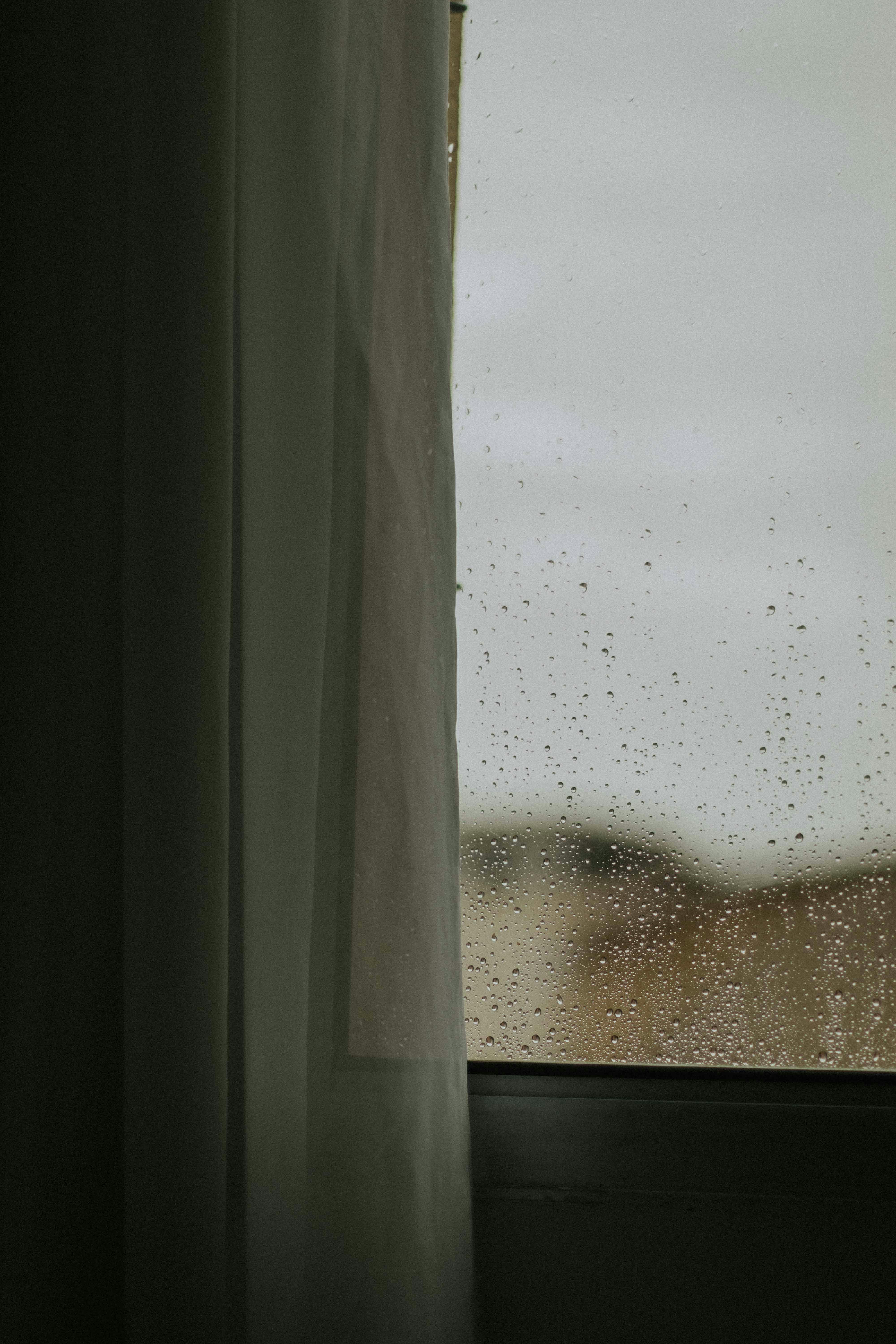 Free A tranquil scene of rain on a window with sheer curtains flowing gently inside. Stock Photo