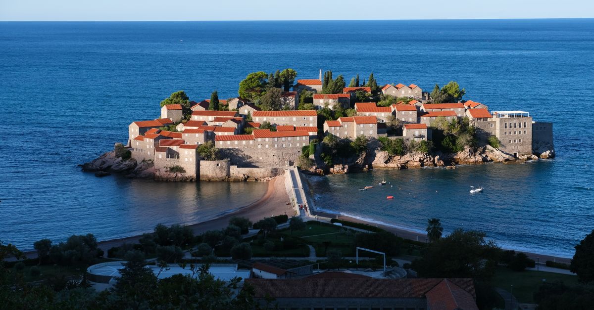 A plane flies over the rugged, green mountains and historic coastal red-roofed buildings of Montenegro's bay.