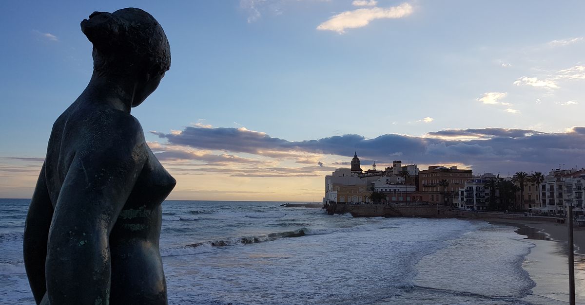 A serene view of the Sitges coastline with a statue in the foreground during sunset.