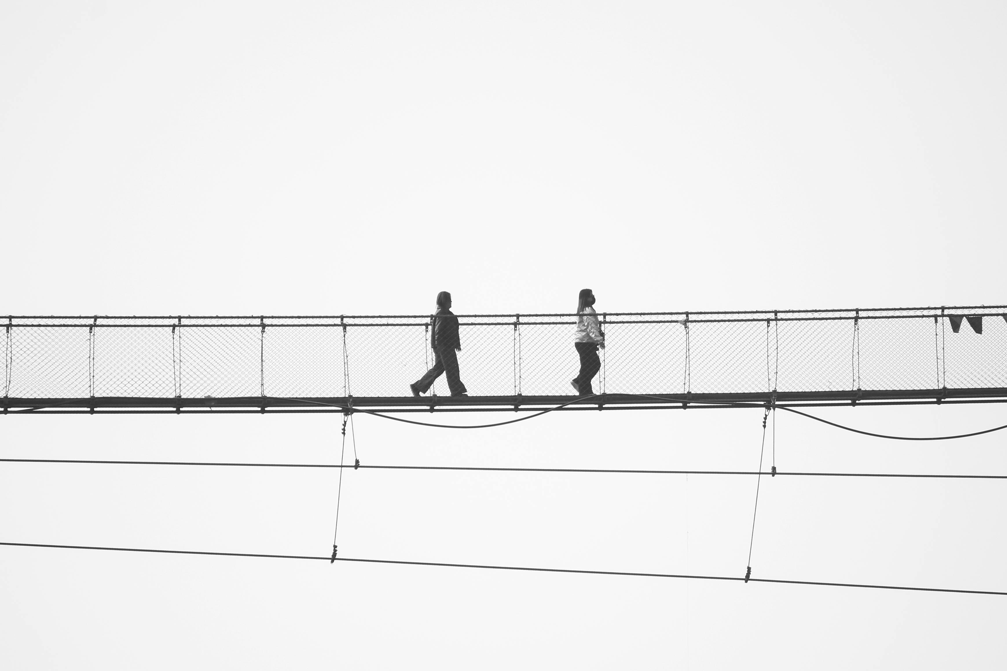 Kostenlos Ein Schwarz-Weiß-Foto von zwei Personen, die auf einer Hängebrücke spazieren gehen und sich als Silhouette vor dem hellen Himmel abheben. Stock-Foto