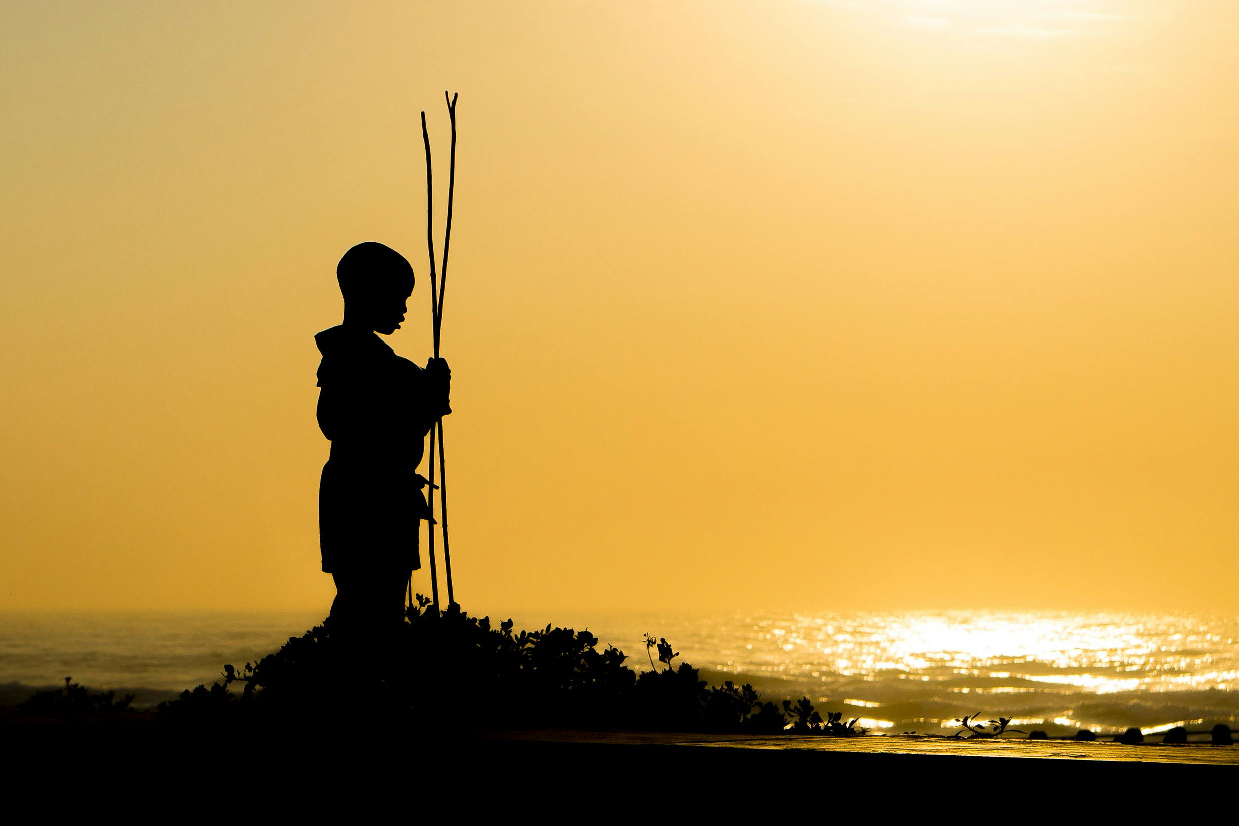 Free A silhouette of a boy holding sticks by the sea during a golden sunset, evoking tranquility. Stock Photo