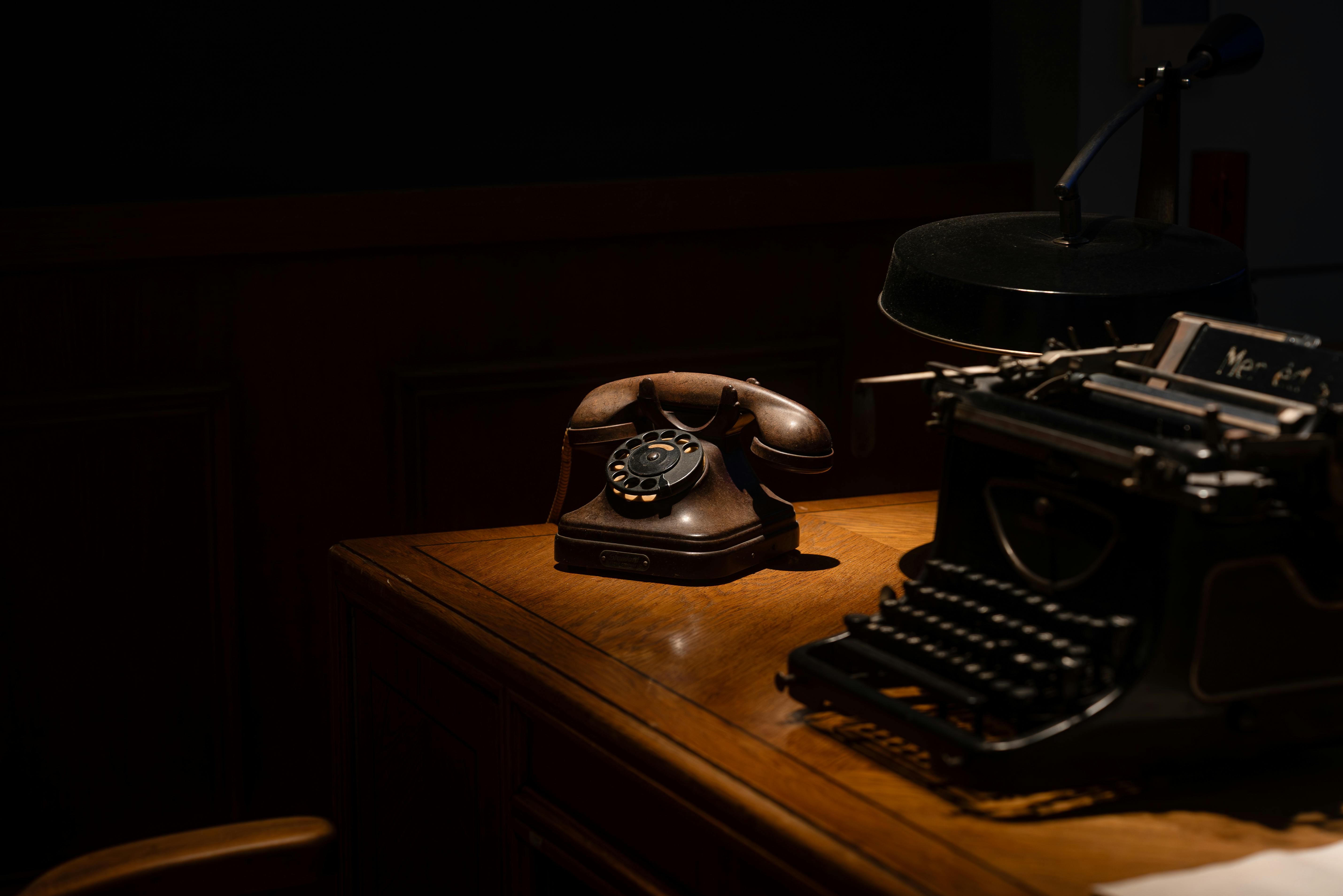 A dimly lit vintage office desk with a rotary telephone and typewriter