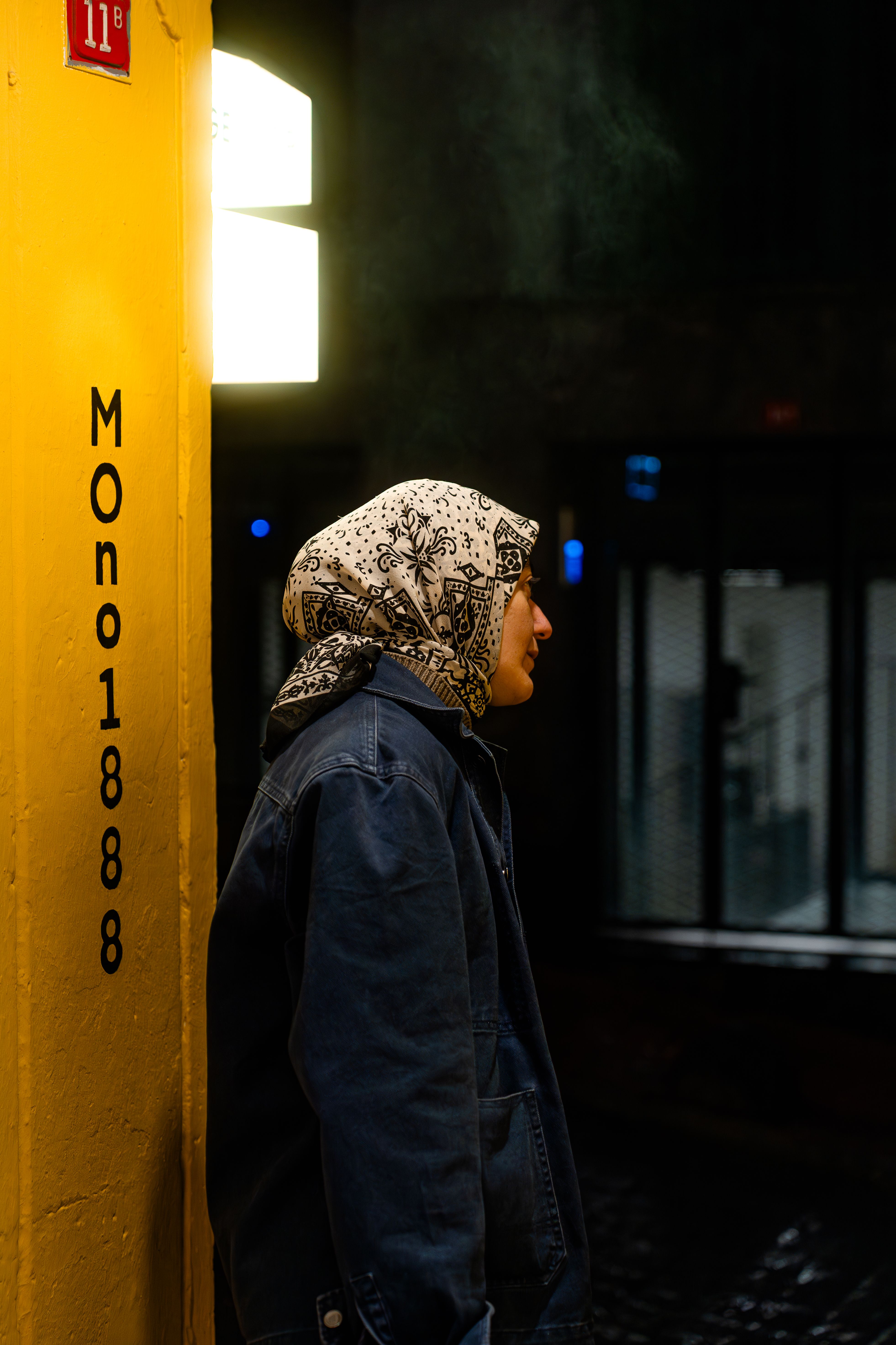 A woman in a headscarf stands by a yellow wall at night, illuminated by a light.