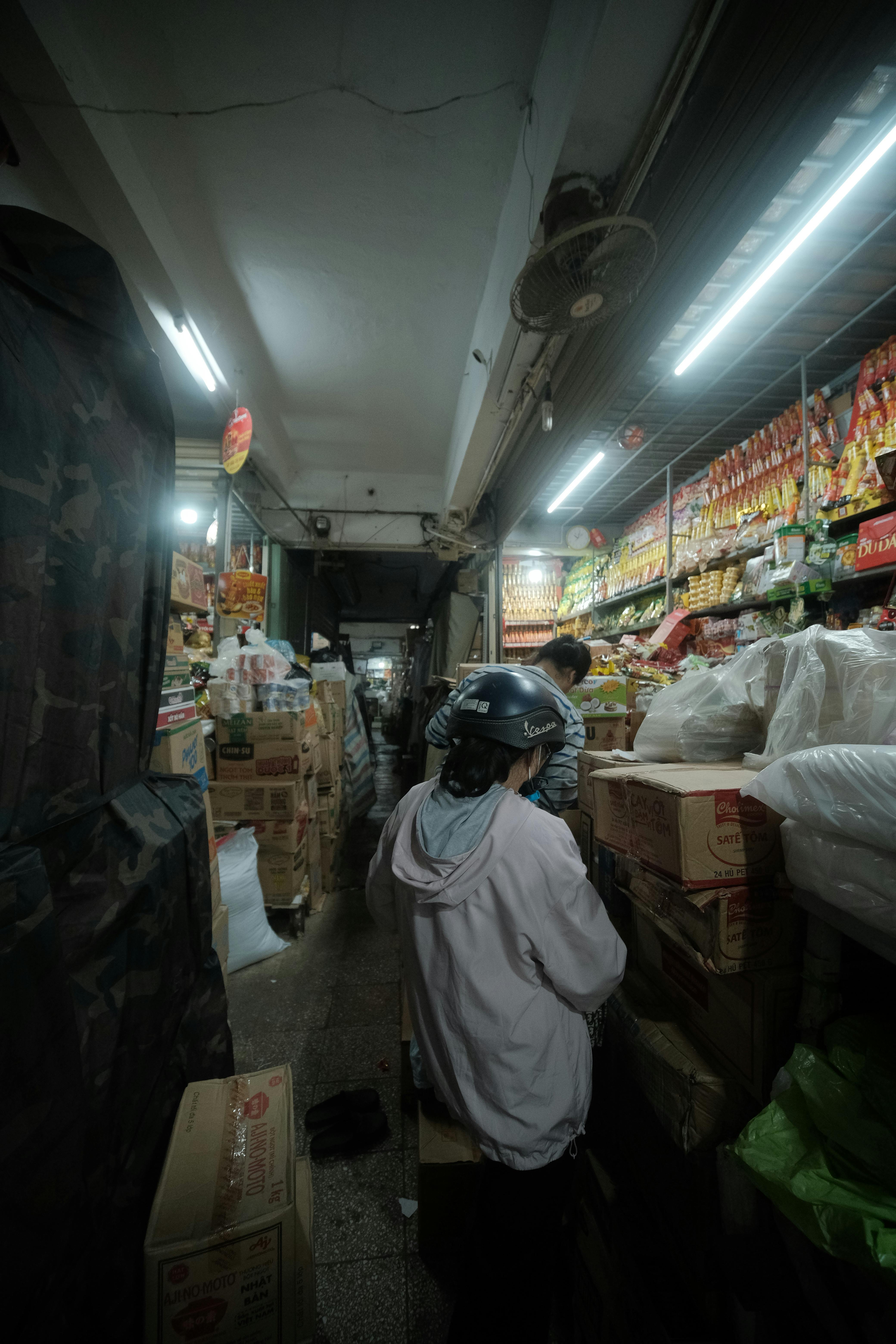 Indoor market scene with people shopping in a narrow aisle filled with goods.