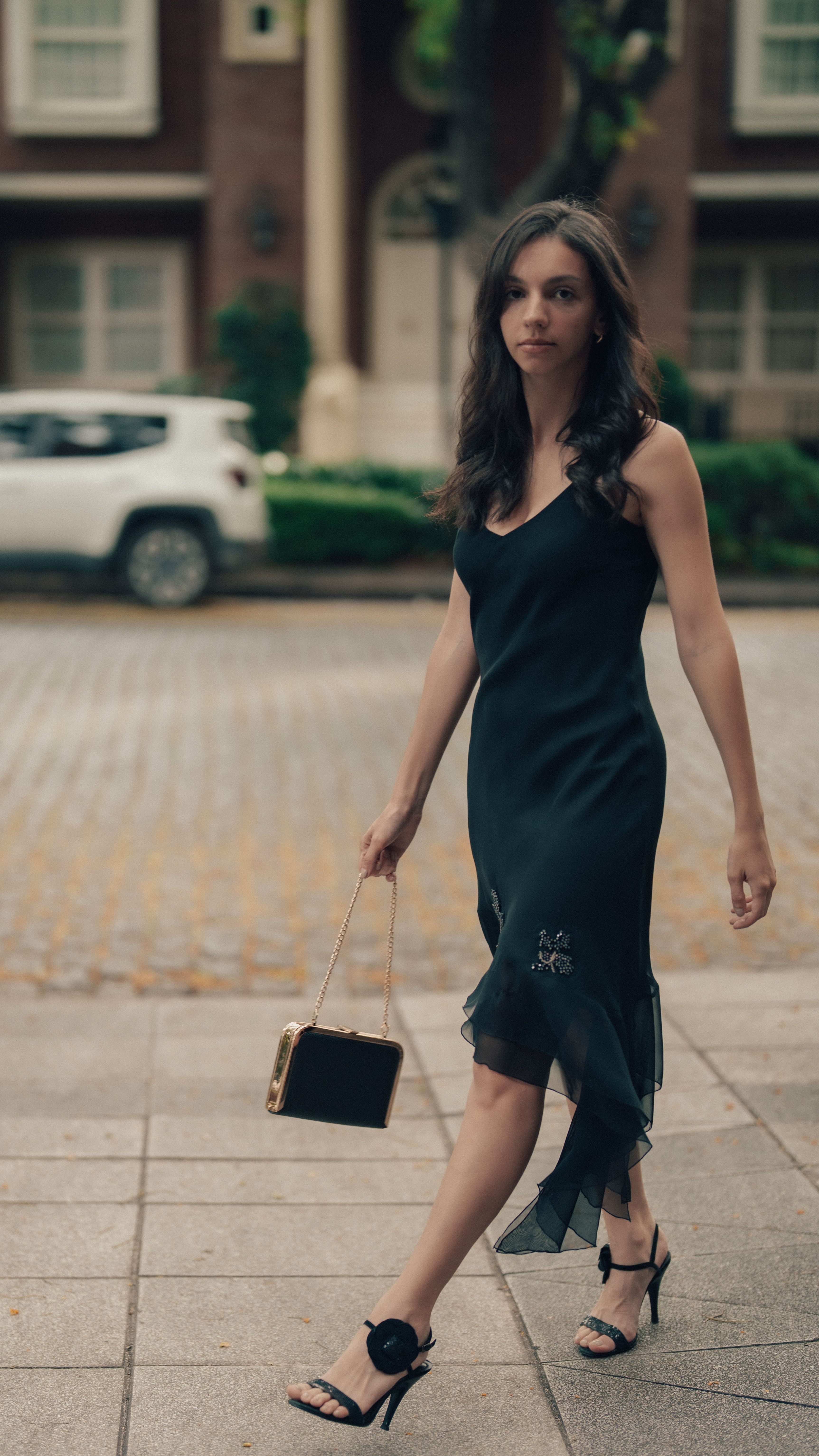 Woman in black dress walking on city street, holding handbag.