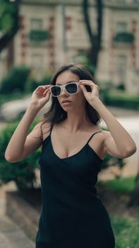 Elegant woman in a black dress and sunglasses posing outdoors on a summer day.