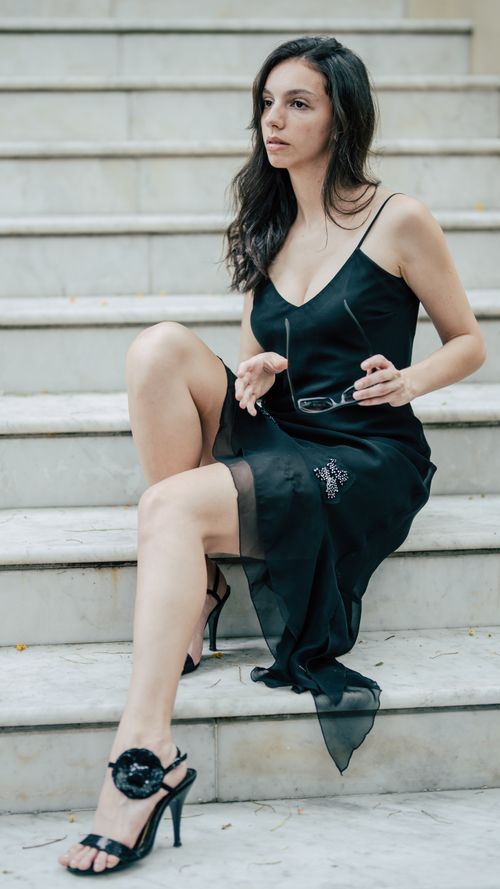Free Chic woman in a black dress sitting thoughtfully on marble stairs holding glasses. Stock Photo