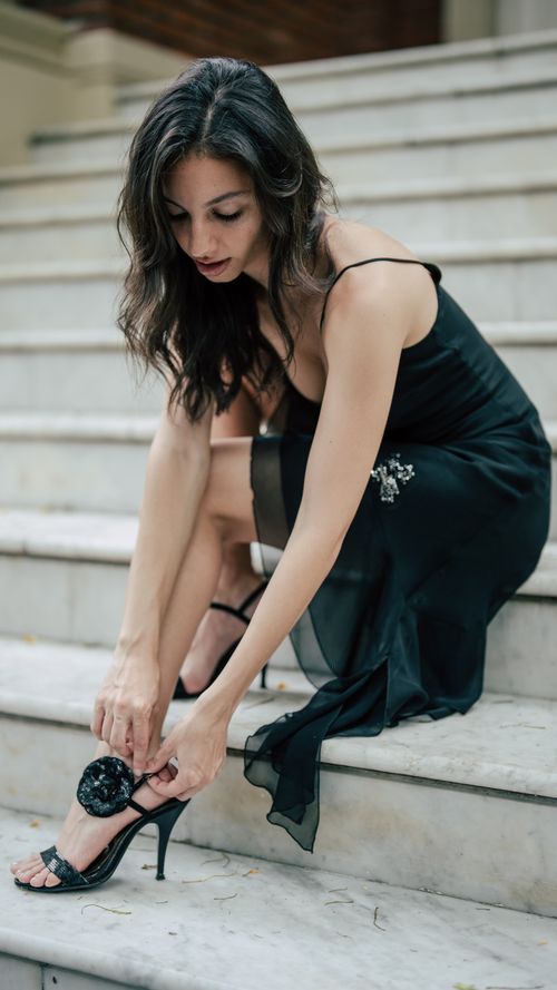 Free Woman in black dress adjusting heels, seated on stairs. Elegant evening fashion. Stock Photo