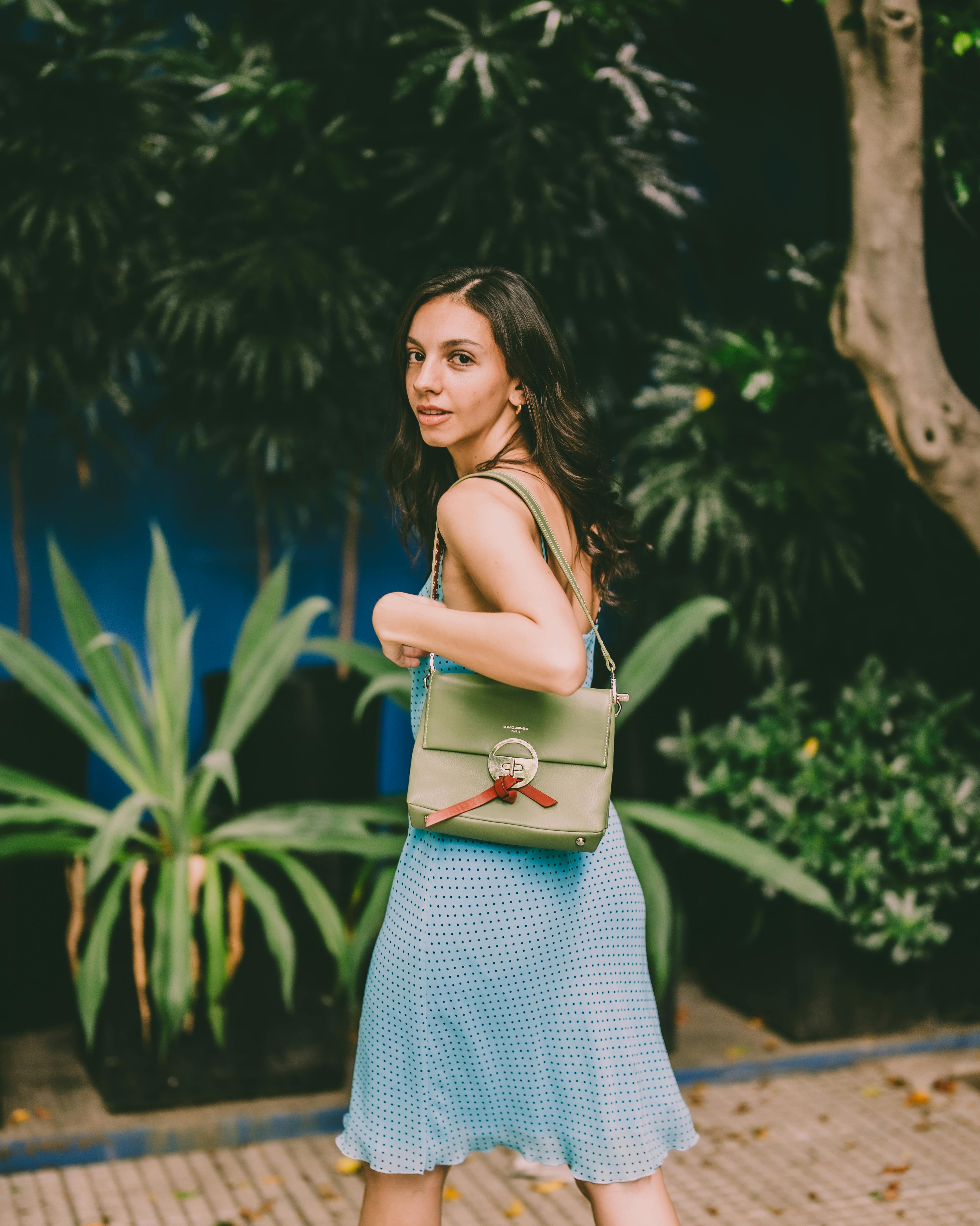 Free Young woman in blue dress carrying a green handbag in a lush outdoor setting. Stock Photo