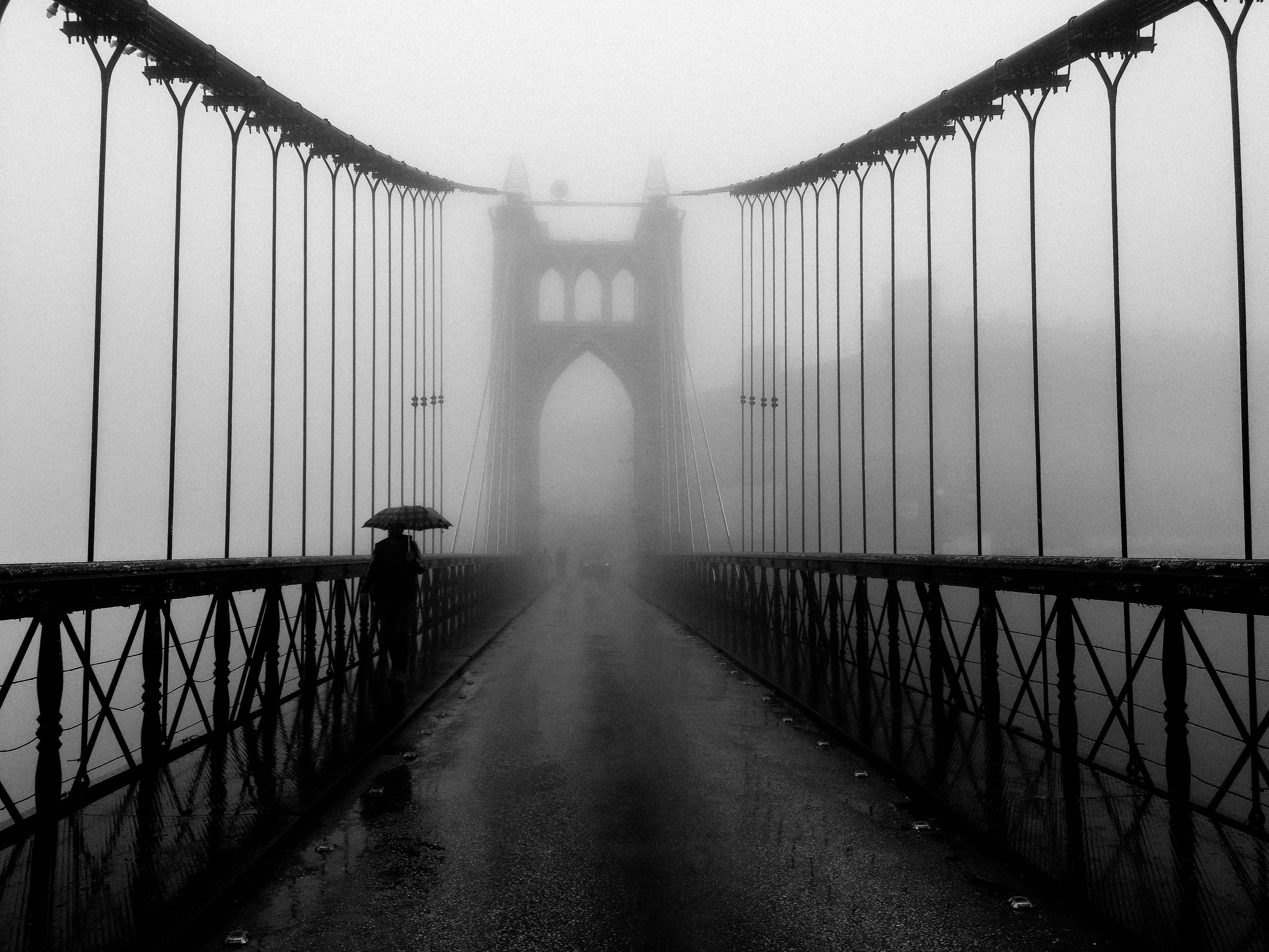 A black and white photo of a person walking on a foggy suspension bridge with an umbrella.