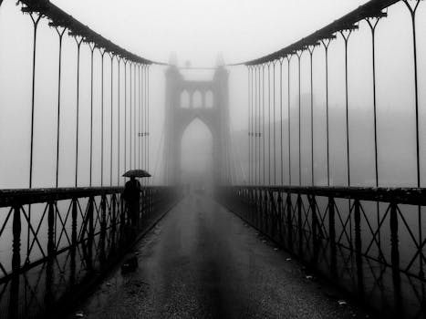 A black and white photo of a person walking on a foggy suspension bridge with an umbrella.