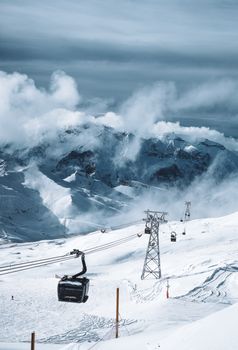 Dramatic view of cable cars crossing snowy mountain peaks under cloudy skies.