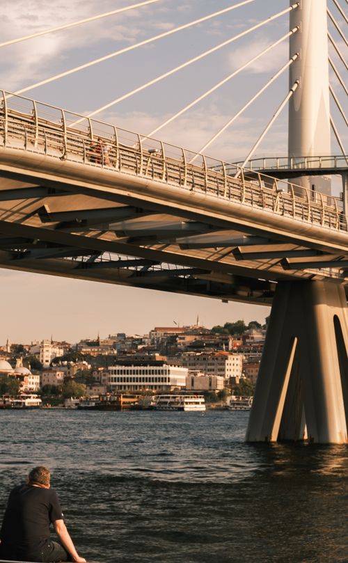 Free Urban bridge in cityscape with waterfront view and a person relaxing by the riverbank. Stock Photo