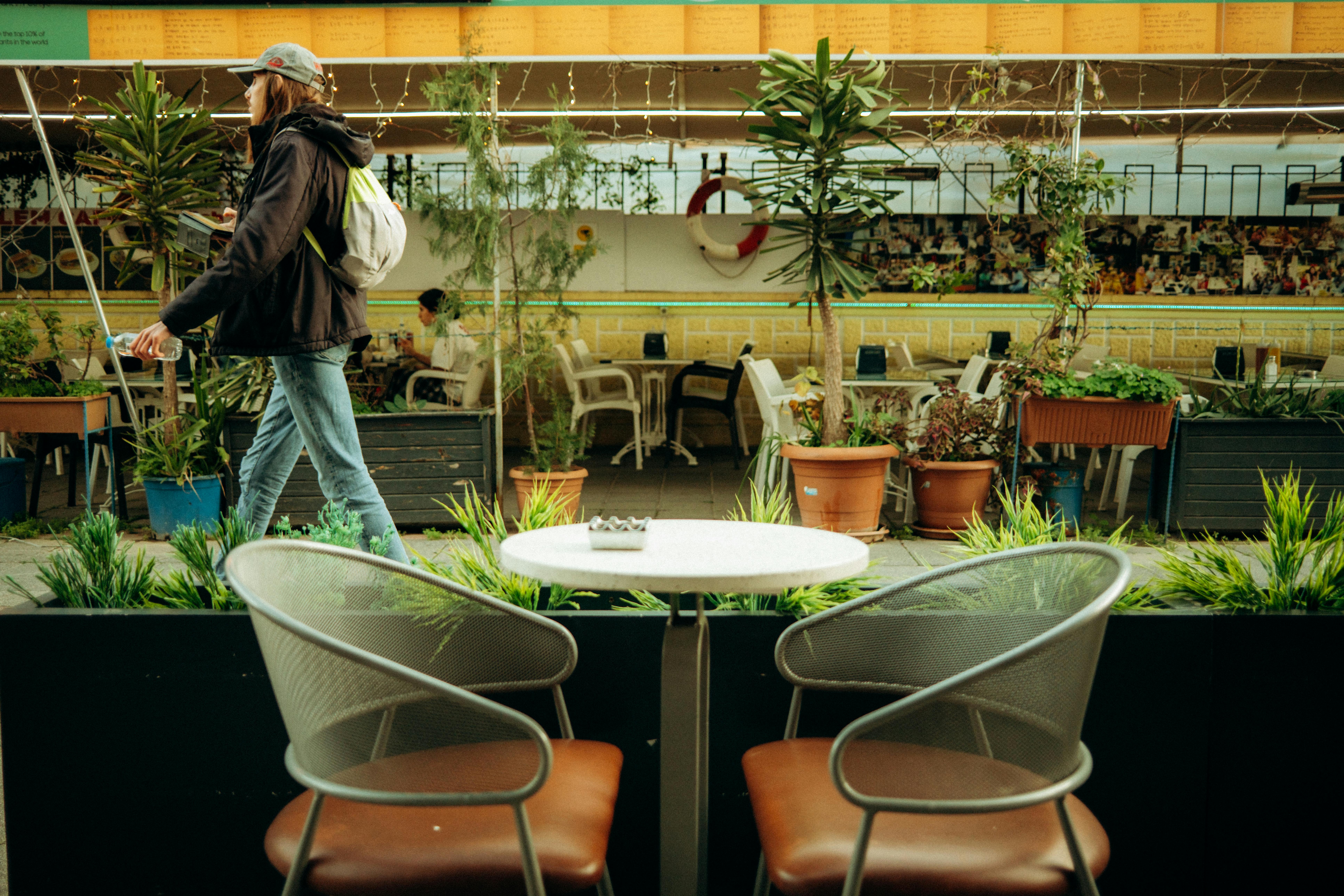 Gratis Vista ambientale di un bar all'aperto con sedie vuote e un passante, che cattura il relax urbano. Foto a disposizione
