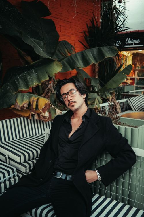 Free Fashionable man in a black suit sitting outdoors on striped cushions, surrounded by lush plants. Stock Photo