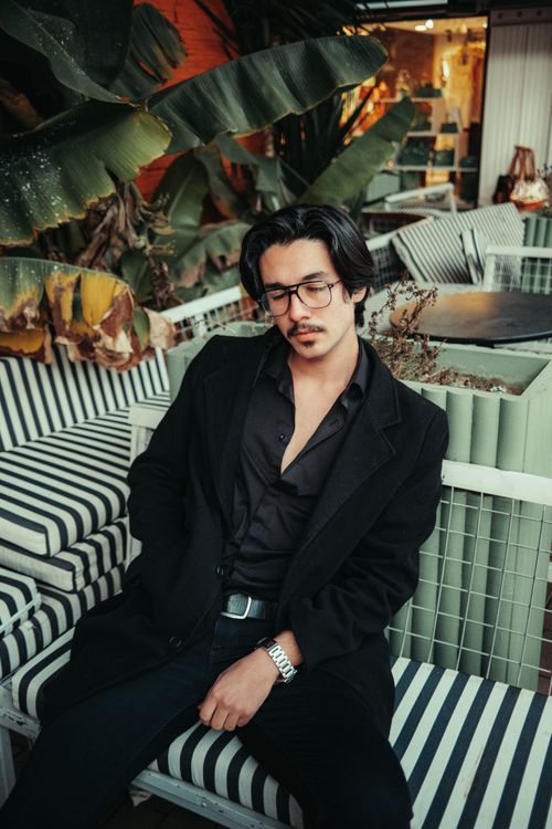 Free A fashionable man in a black outfit relaxes in an outdoor cafe with lush greenery. Stock Photo