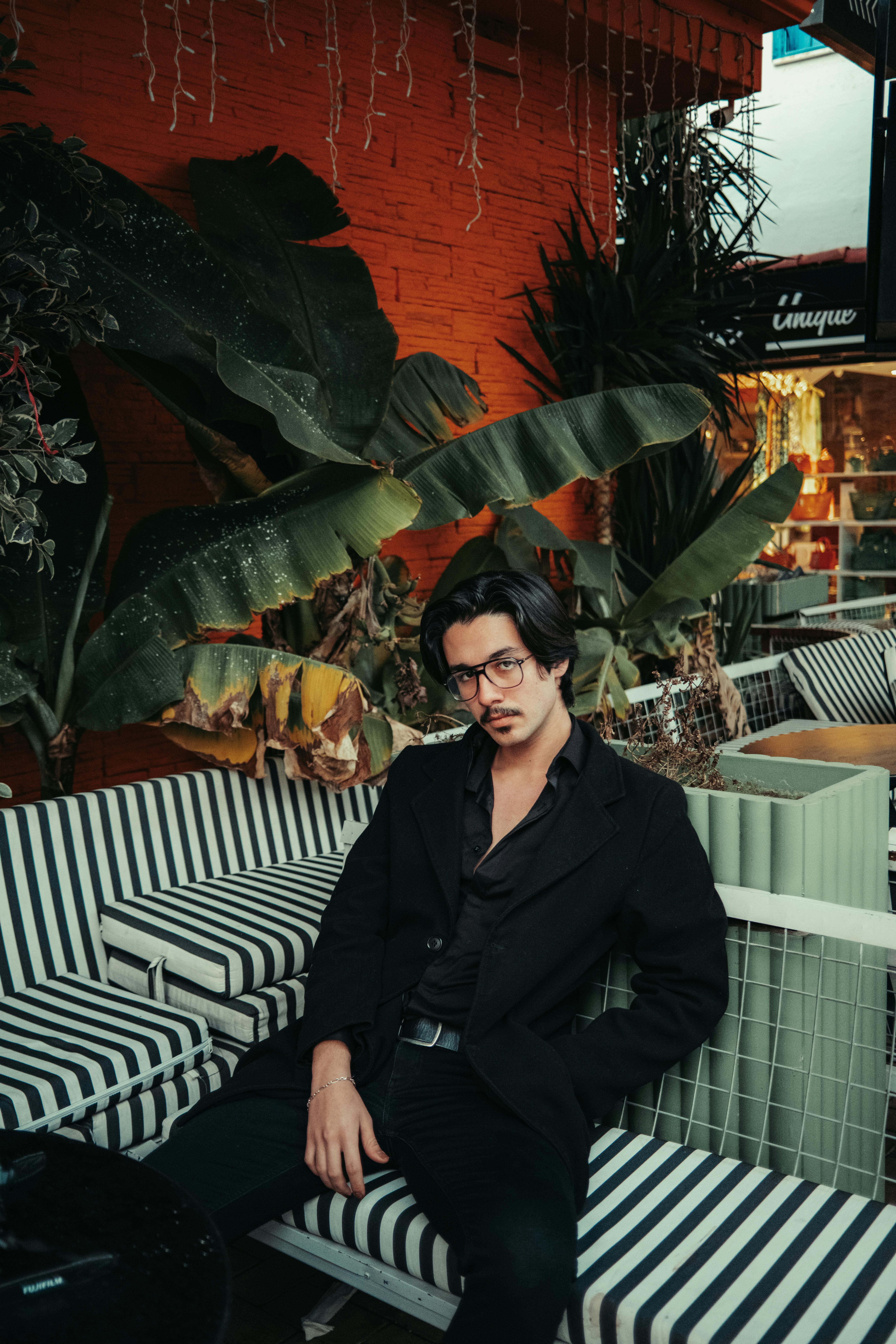 A fashionable man in black clothing sits on a striped sofa outdoors, surrounded by tropical plants.