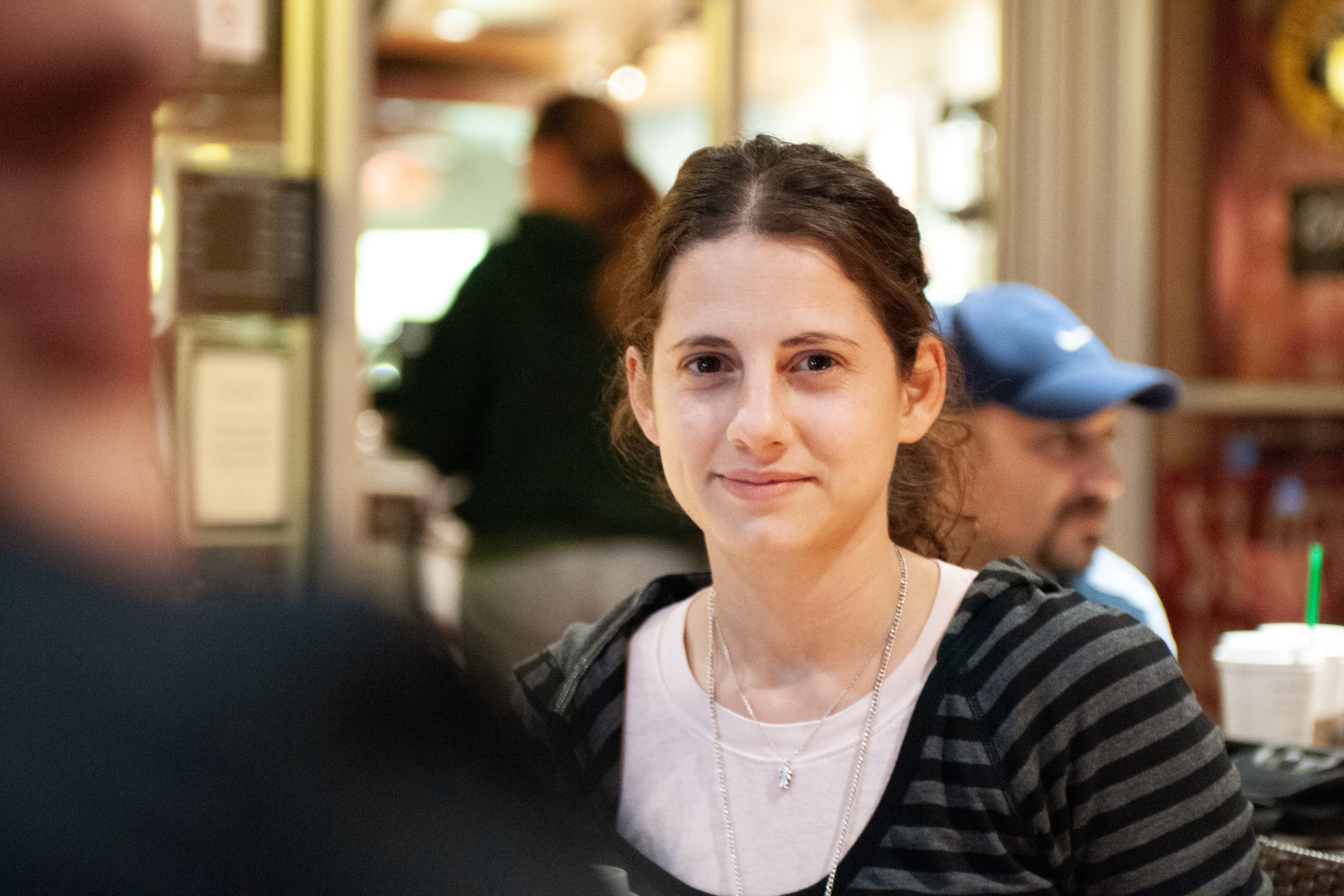 Portrait of a woman in a striped hoodie, sitting in a lively cafe environment.