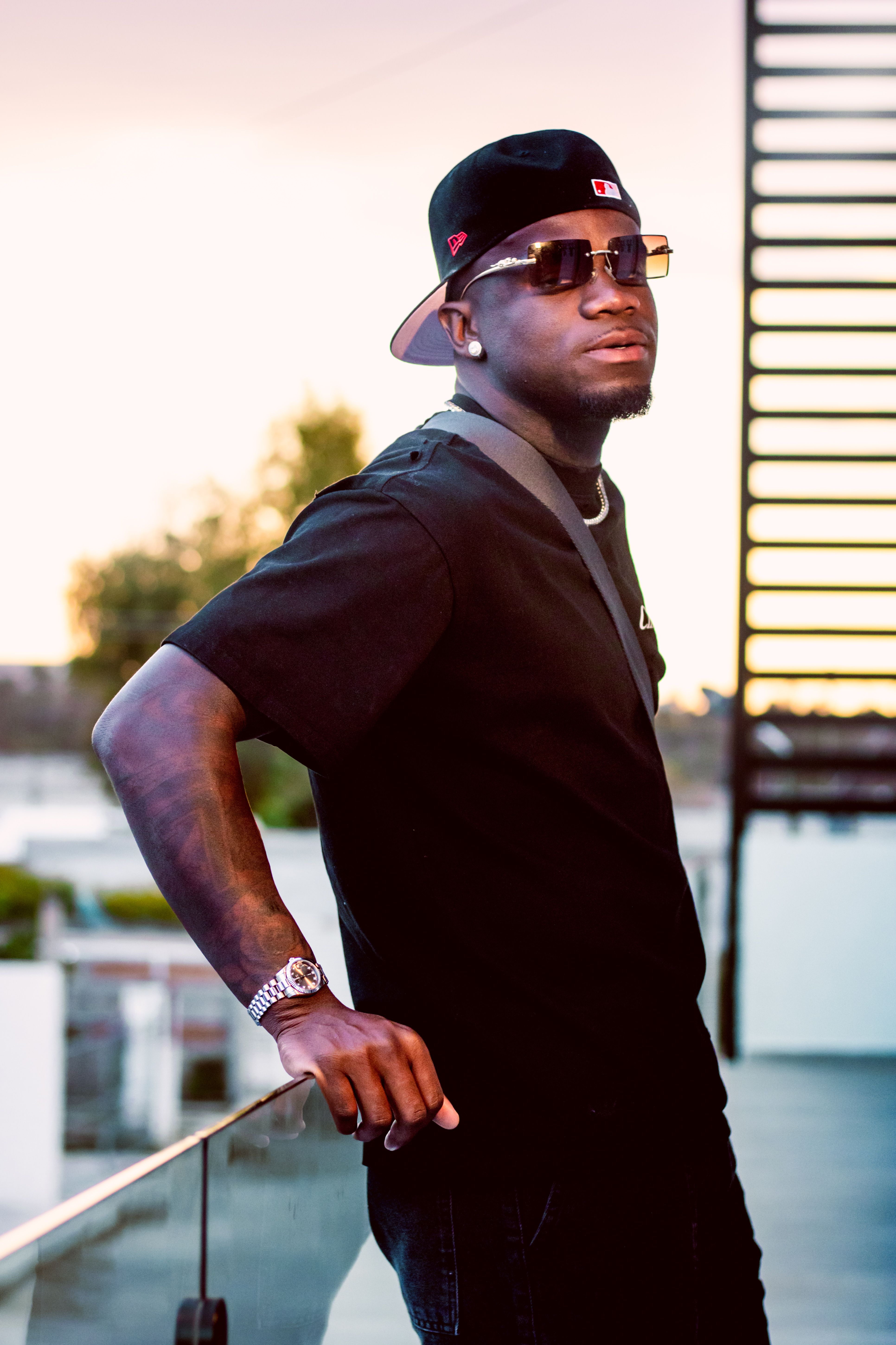 Confident young man wearing sunglasses and cap leans on railing at sunset in urban environment.