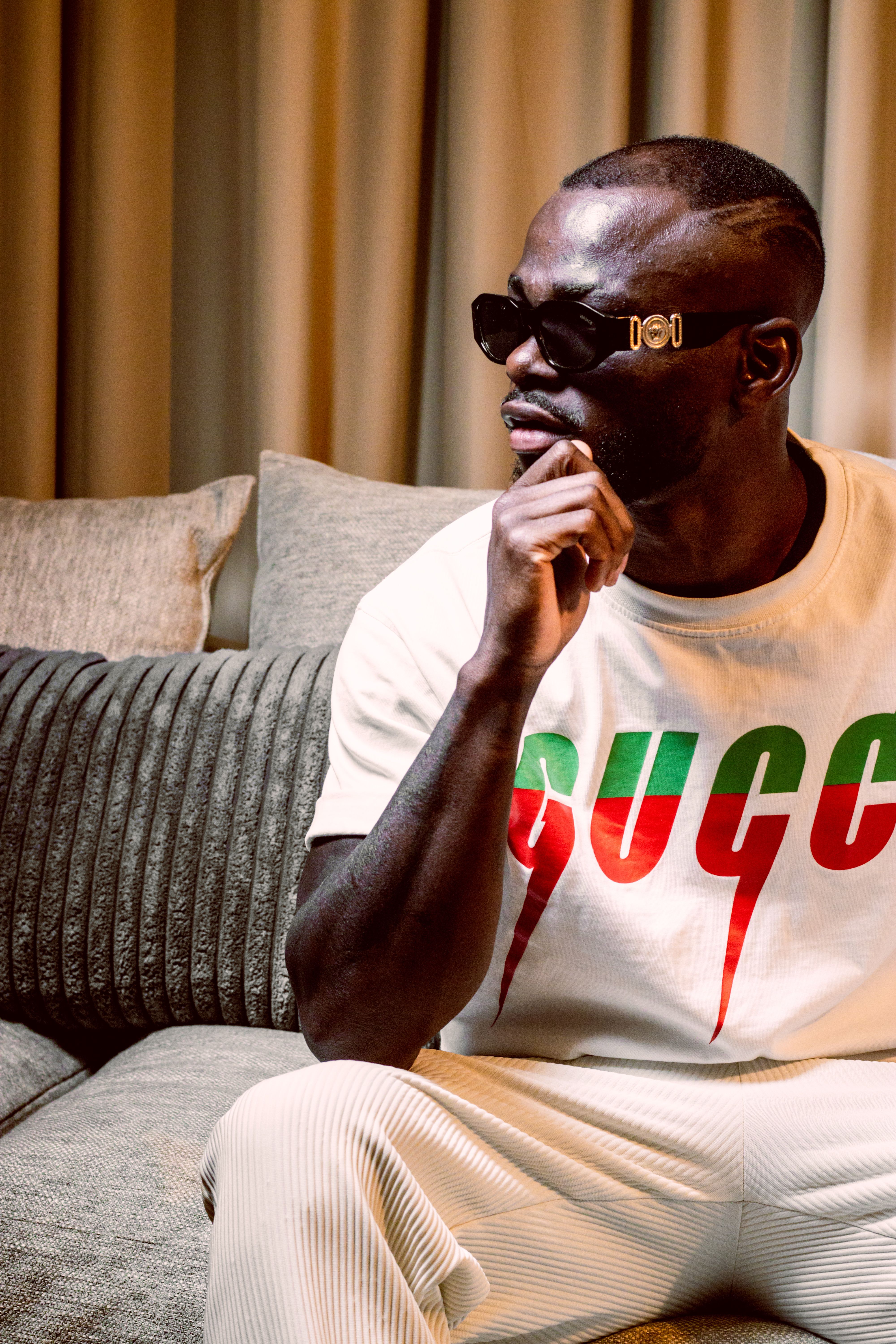 Portrait of a fashionable man sitting on a couch indoors with a reflective pose, wearing sunglasses and casual attire.