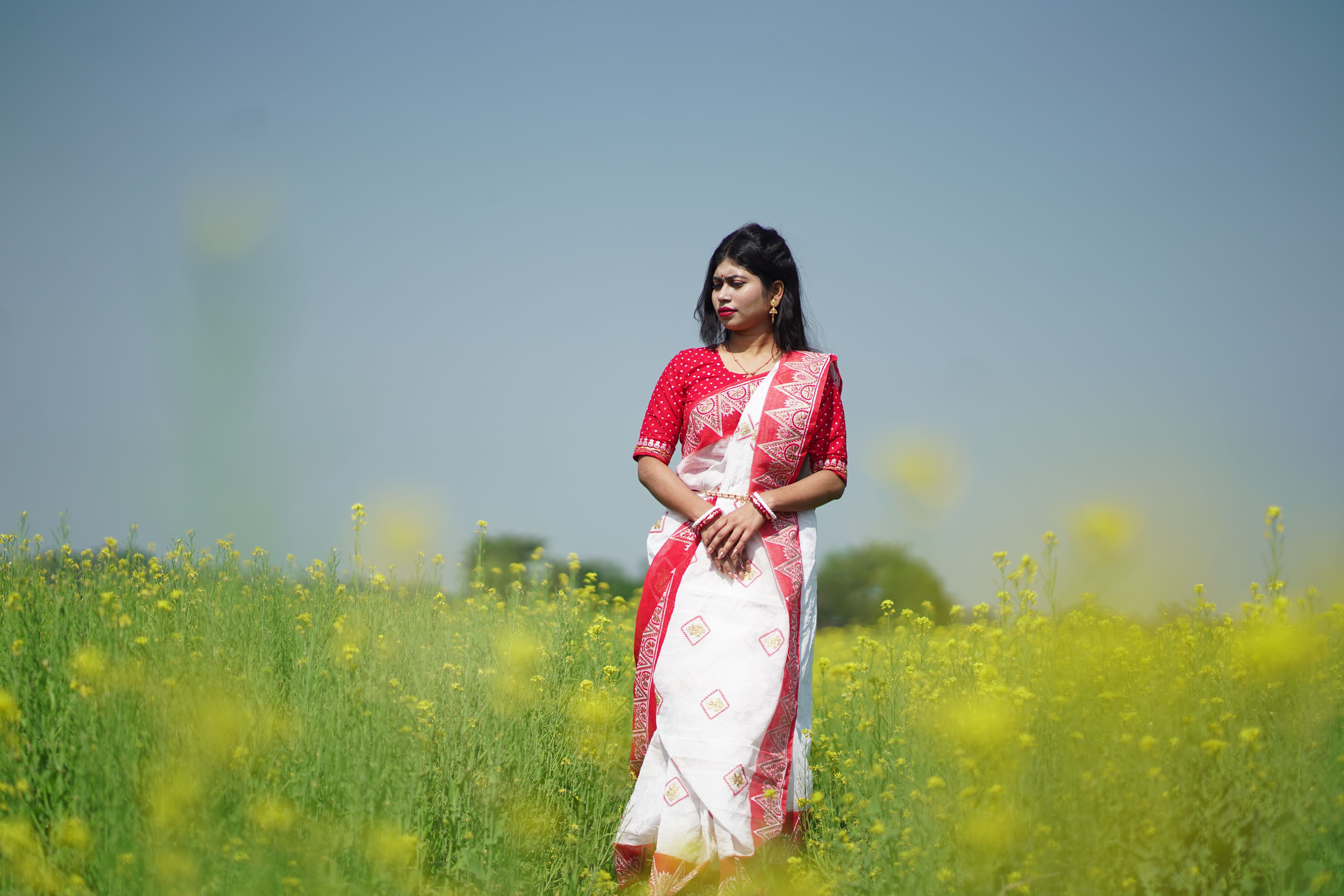 A woman in a red and white saree stands in a vibrant mustard field under clear skies.