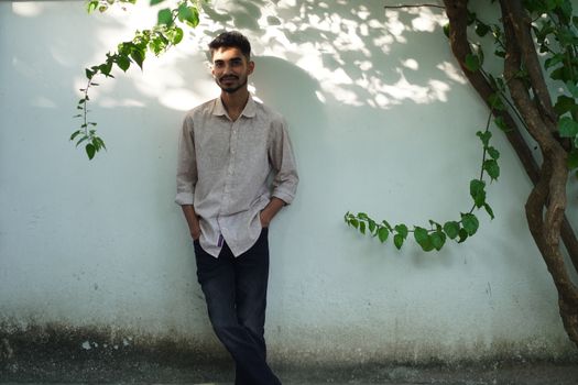 A young man casually leaning against a white wall with greenery.
