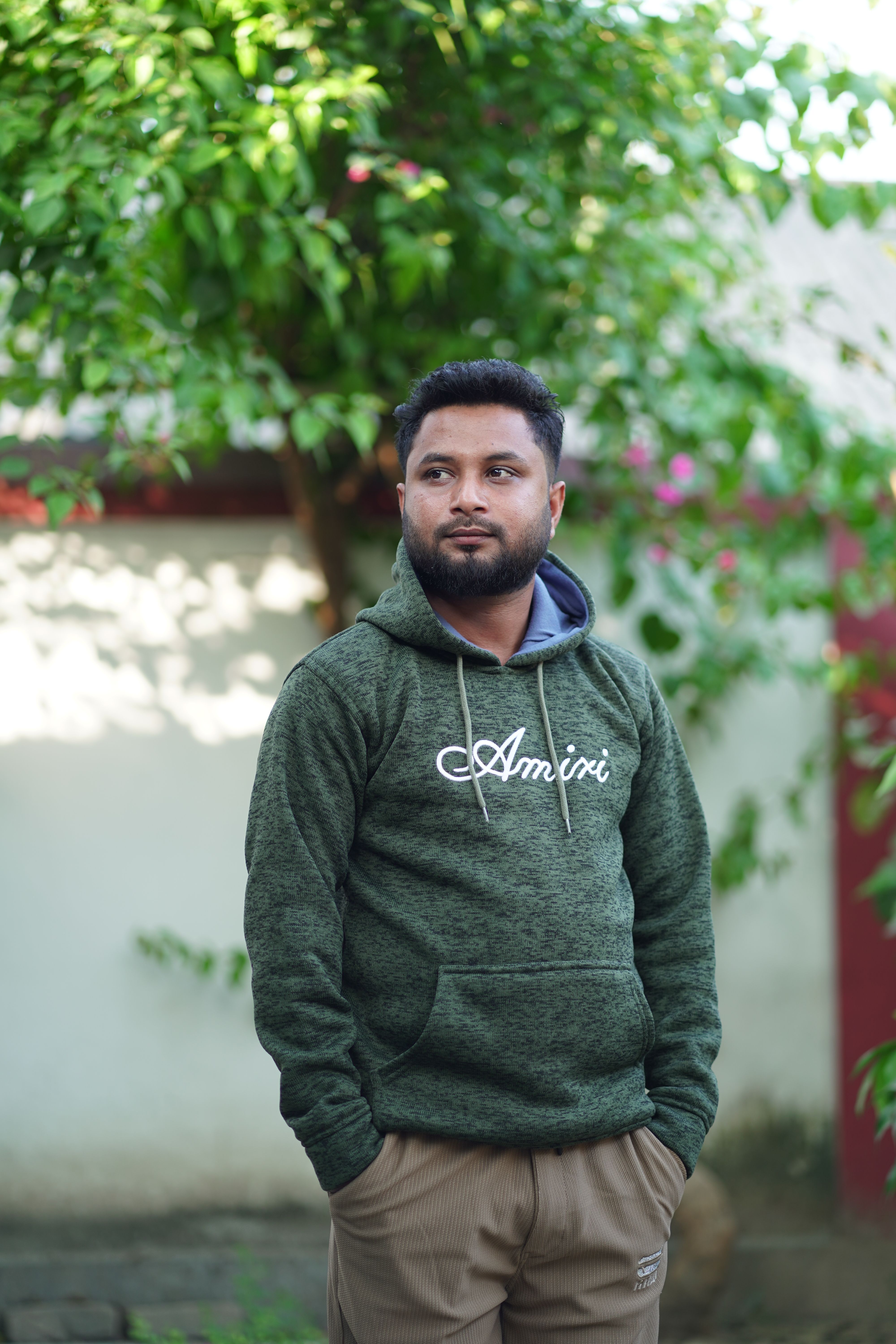 A man wearing a green hoodie stands outdoors near greenery, looking thoughtful.