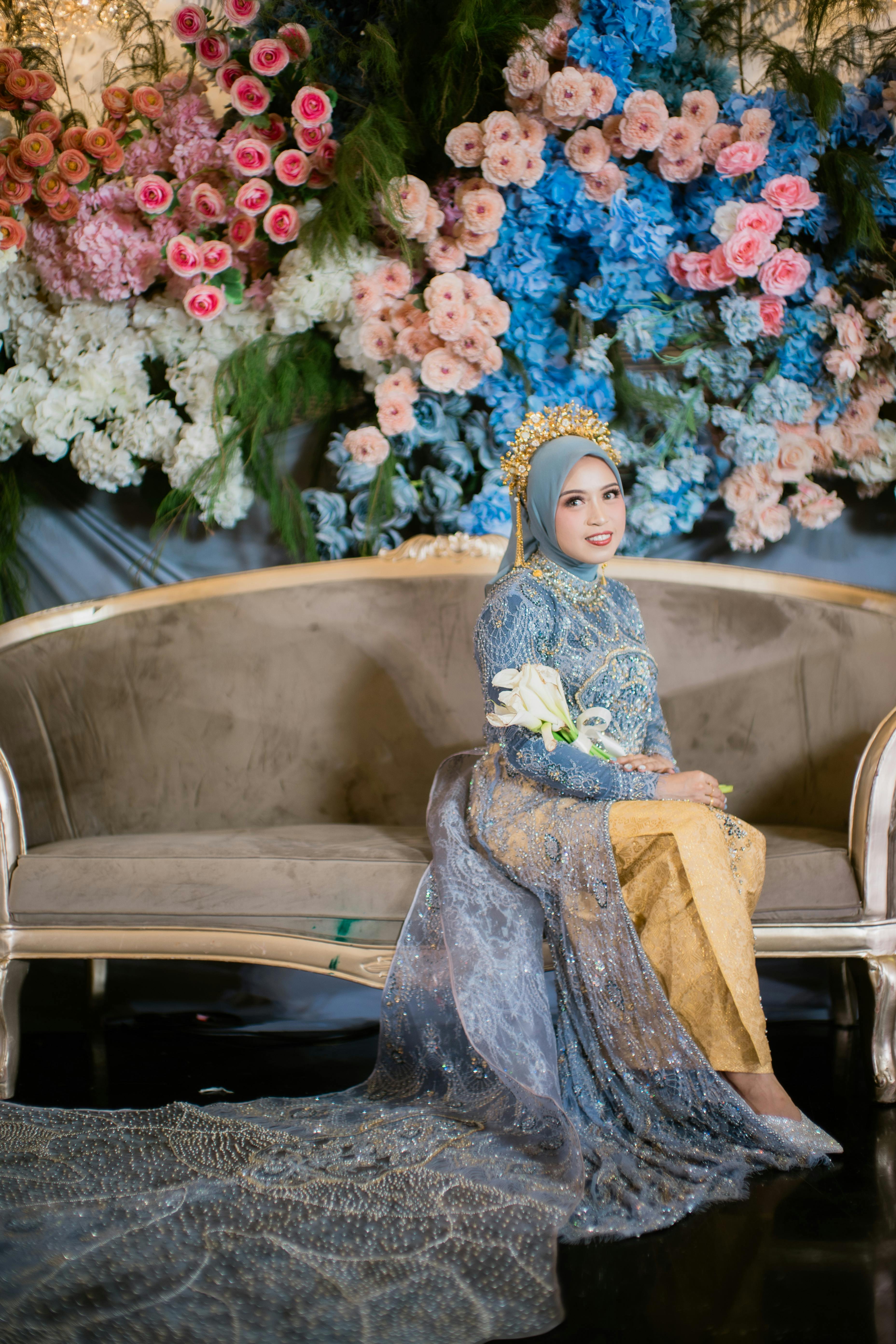 A beautifully dressed bride in traditional attire poses elegantly on a vintage sofa surrounded by colorful flowers.