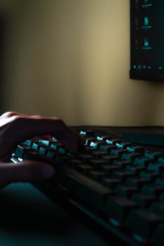 Close-up of a hand on a keyboard with a glowing screen in a dimly lit room.