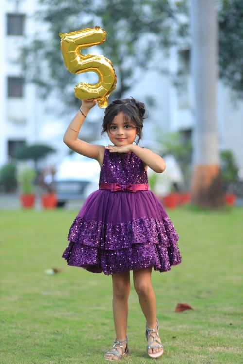 Free A young girl cheerfully holding a number five balloon in a purple dress outdoors. Stock Photo