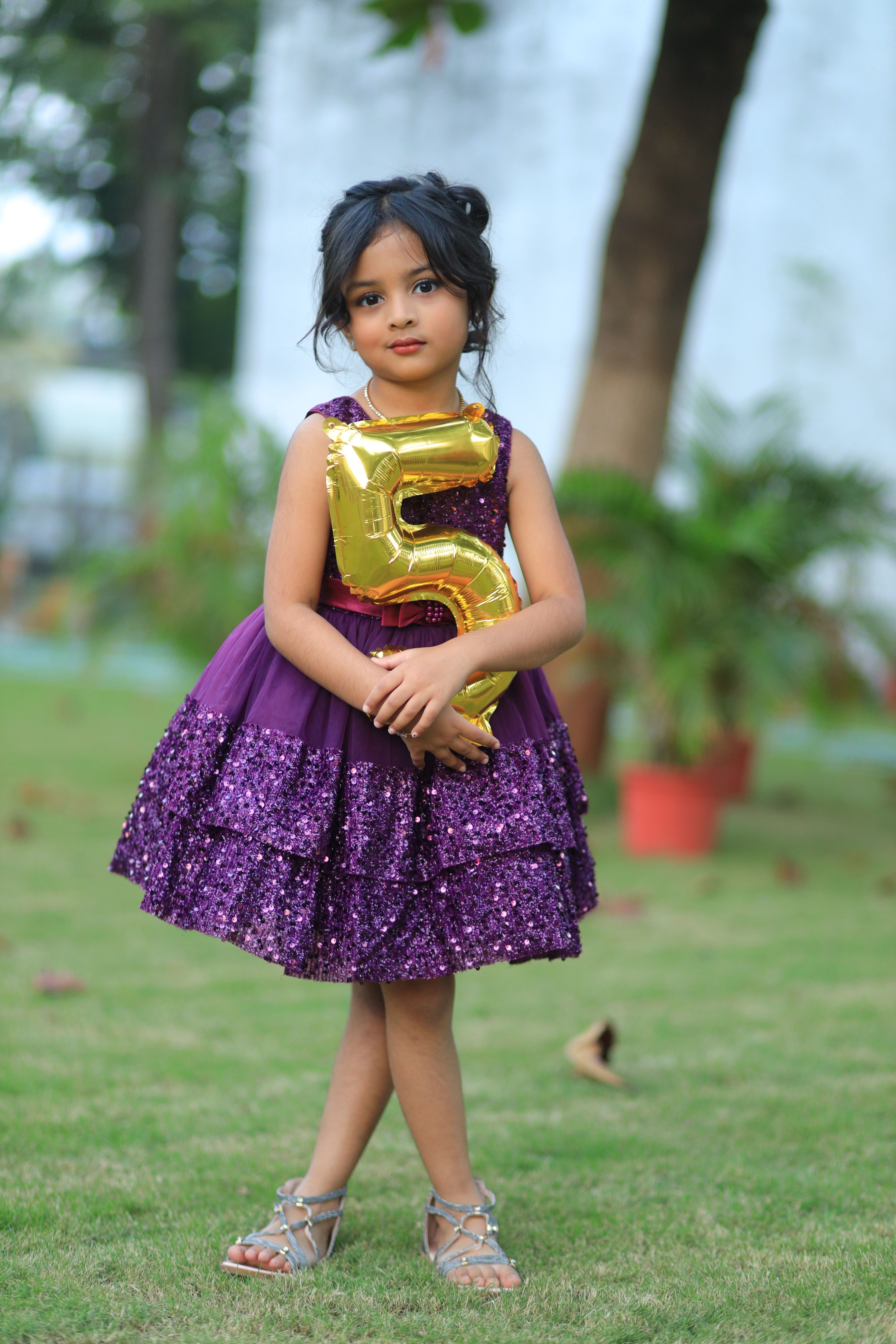 Adorable girl in a glittery purple dress outdoors holding a gold number 5 balloon, perfect for birthday themes.