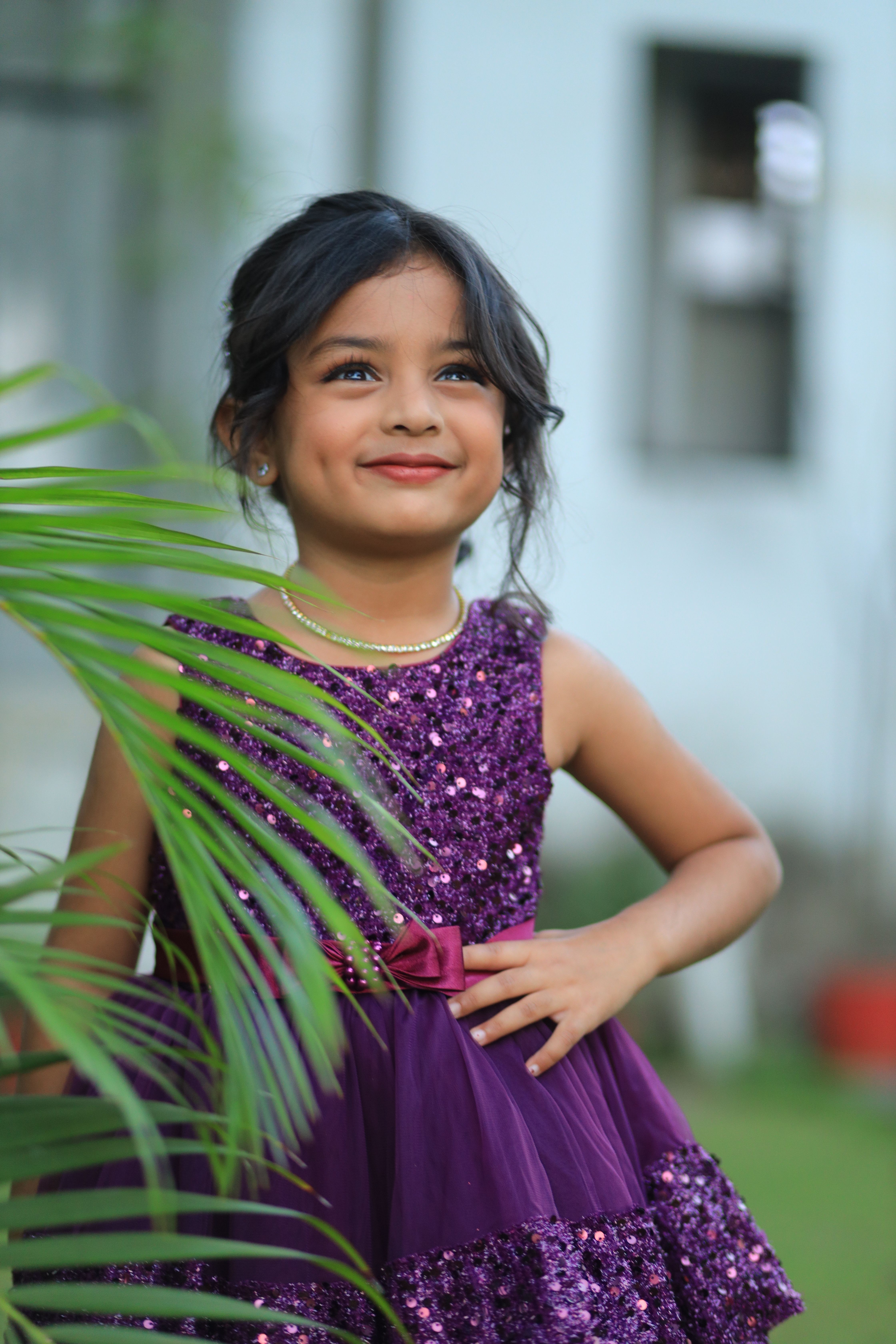 Smiling young girl in a purple dress enjoying a sunny day outdoors among greenery.