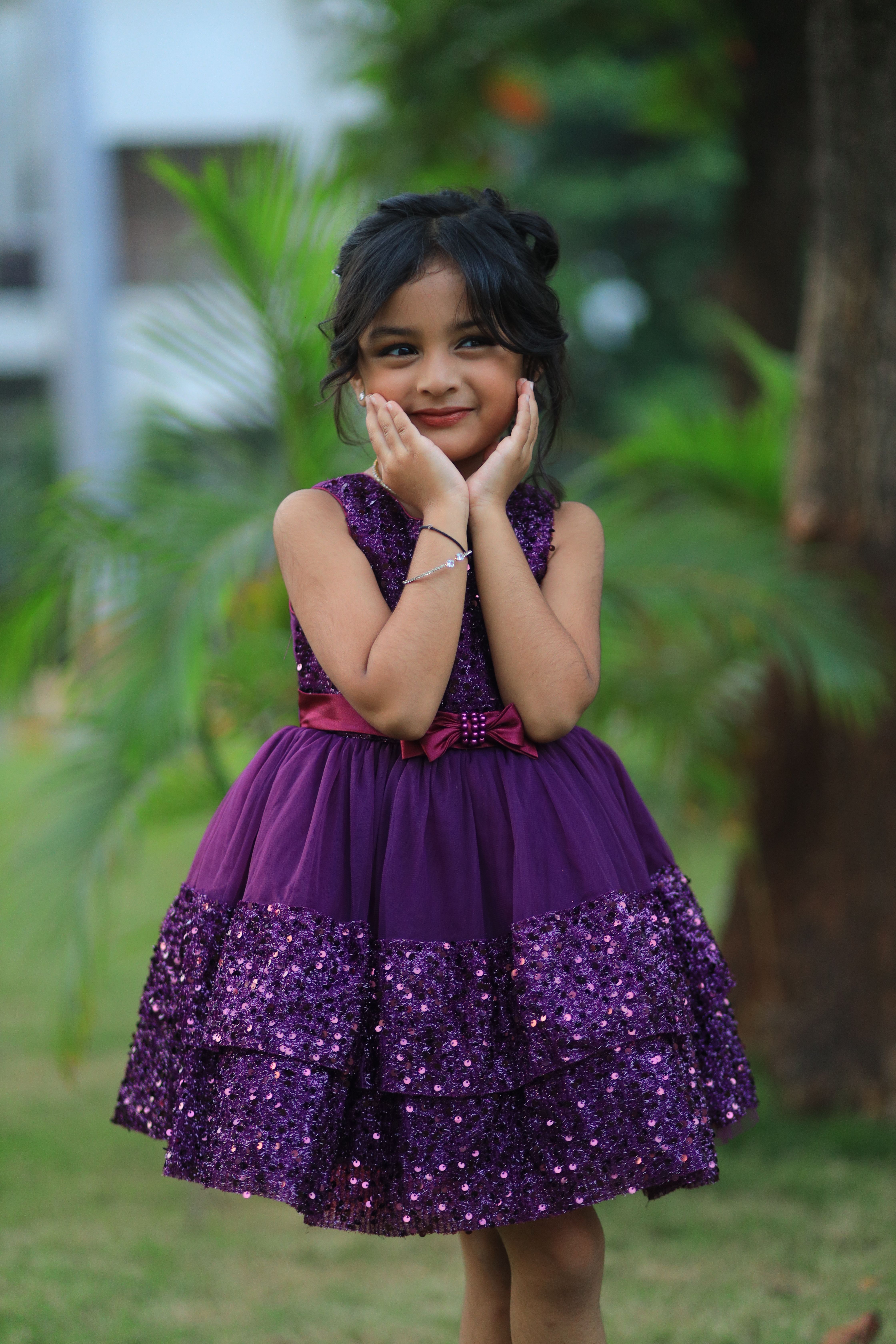 Adorable child in a glittery purple dress posing outside with greenery in the background.
