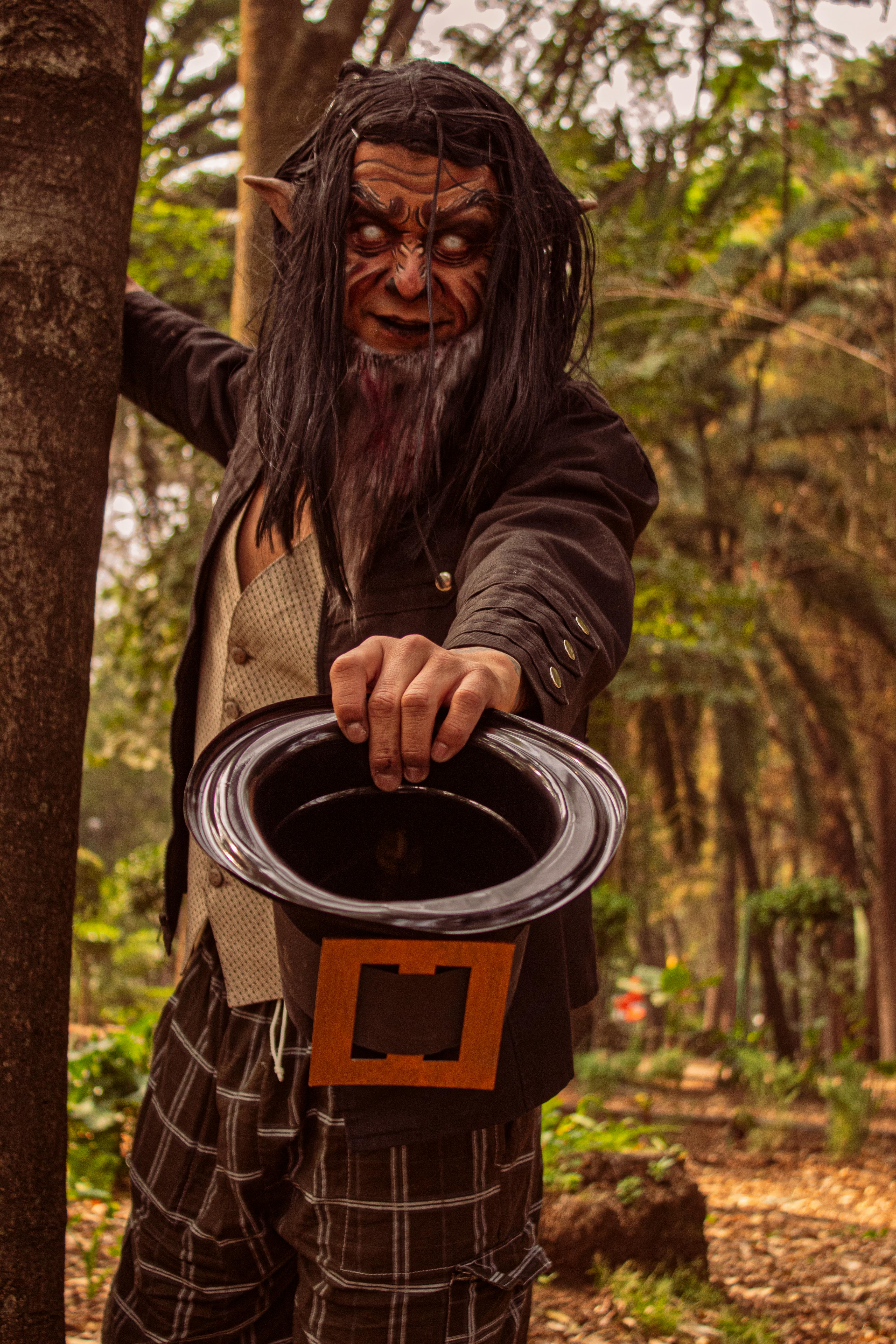 A person dressed as a fantasy creature, holding a top hat in a mystical forest setting.