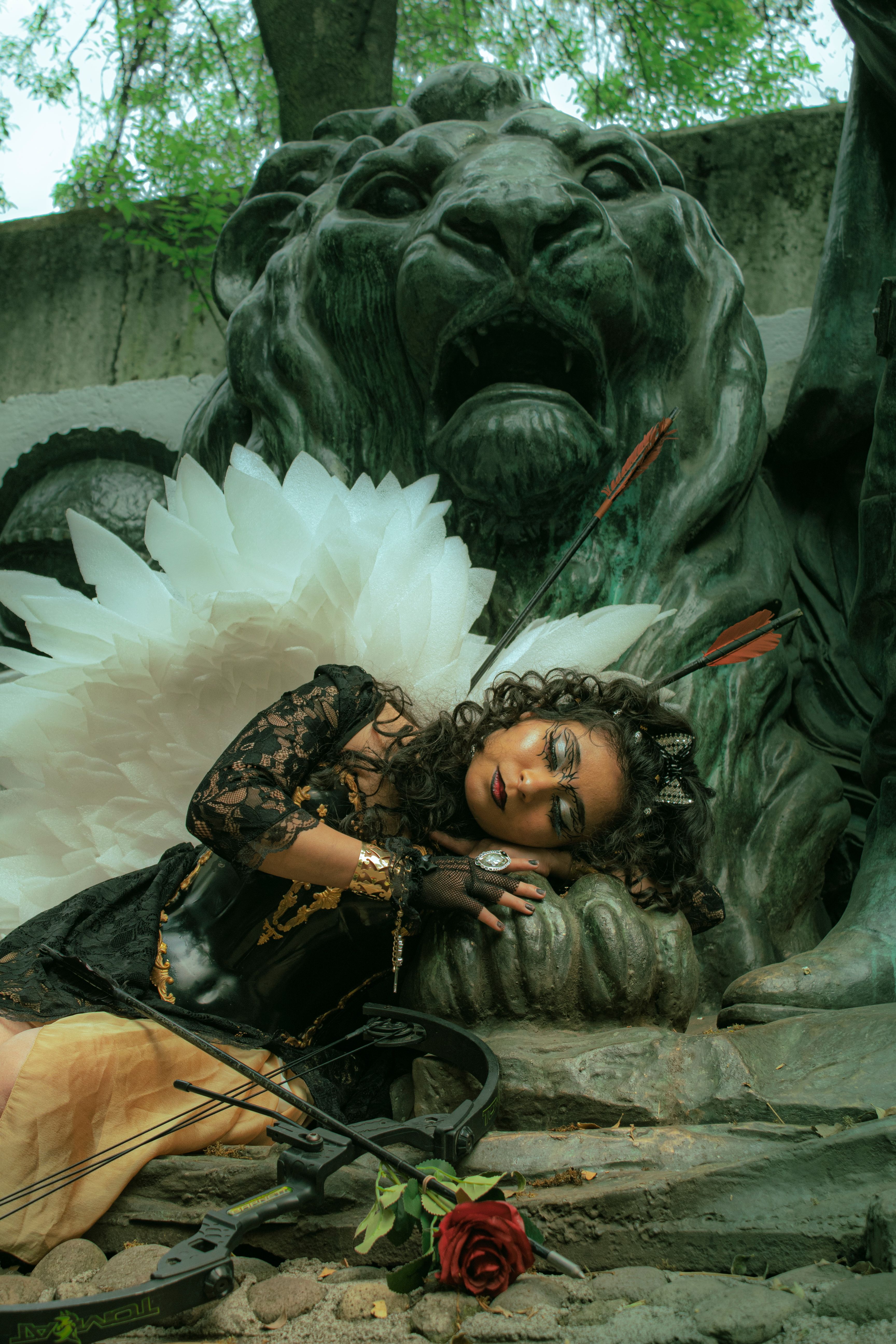 Surreal portrait of an angelic figure with white wings and creative makeup, lying against a lion statue.