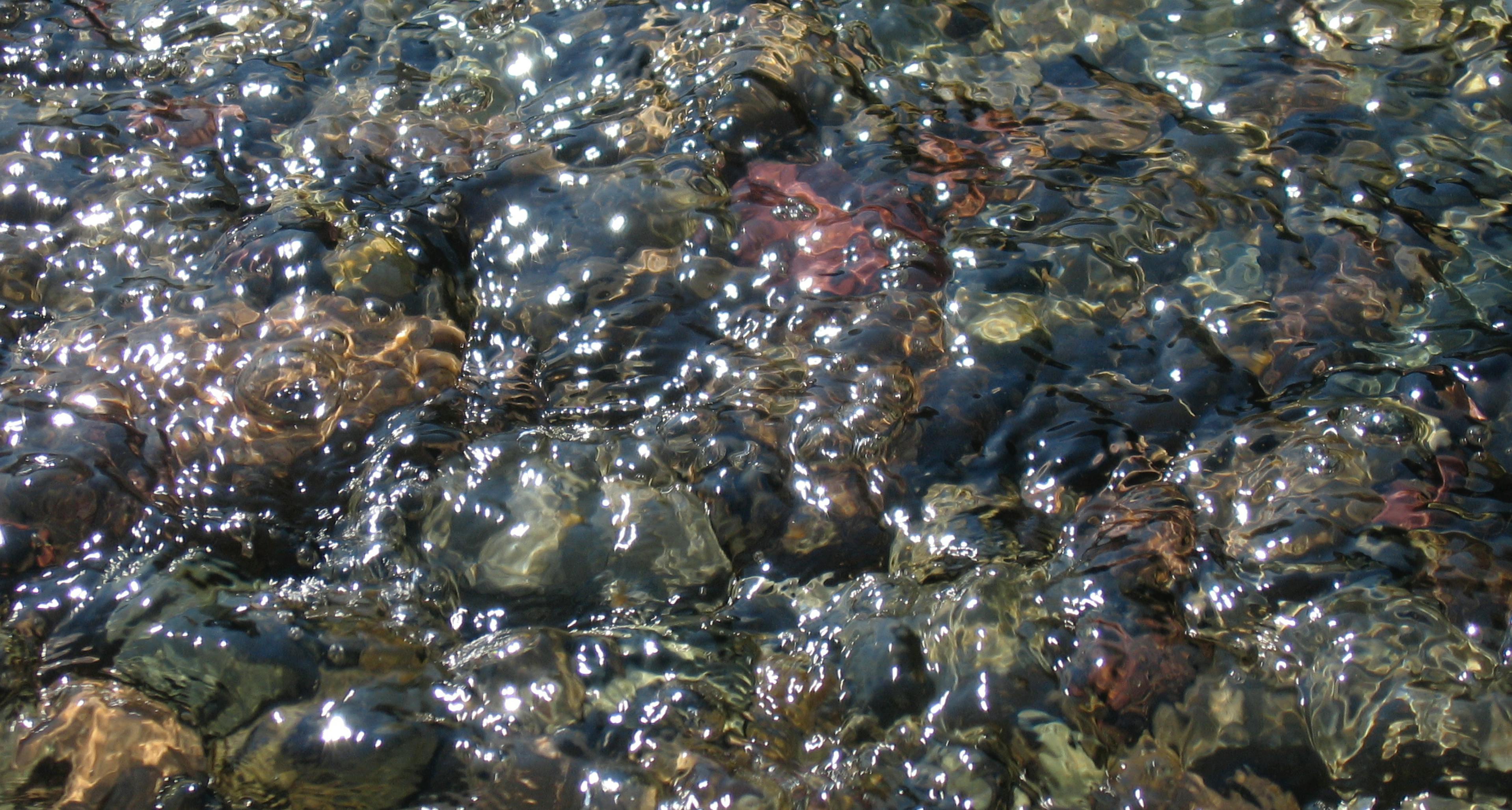 Gratis Primo piano dell'acqua che increspa le pietre colorate, riflettendo la luce del sole. Foto a disposizione